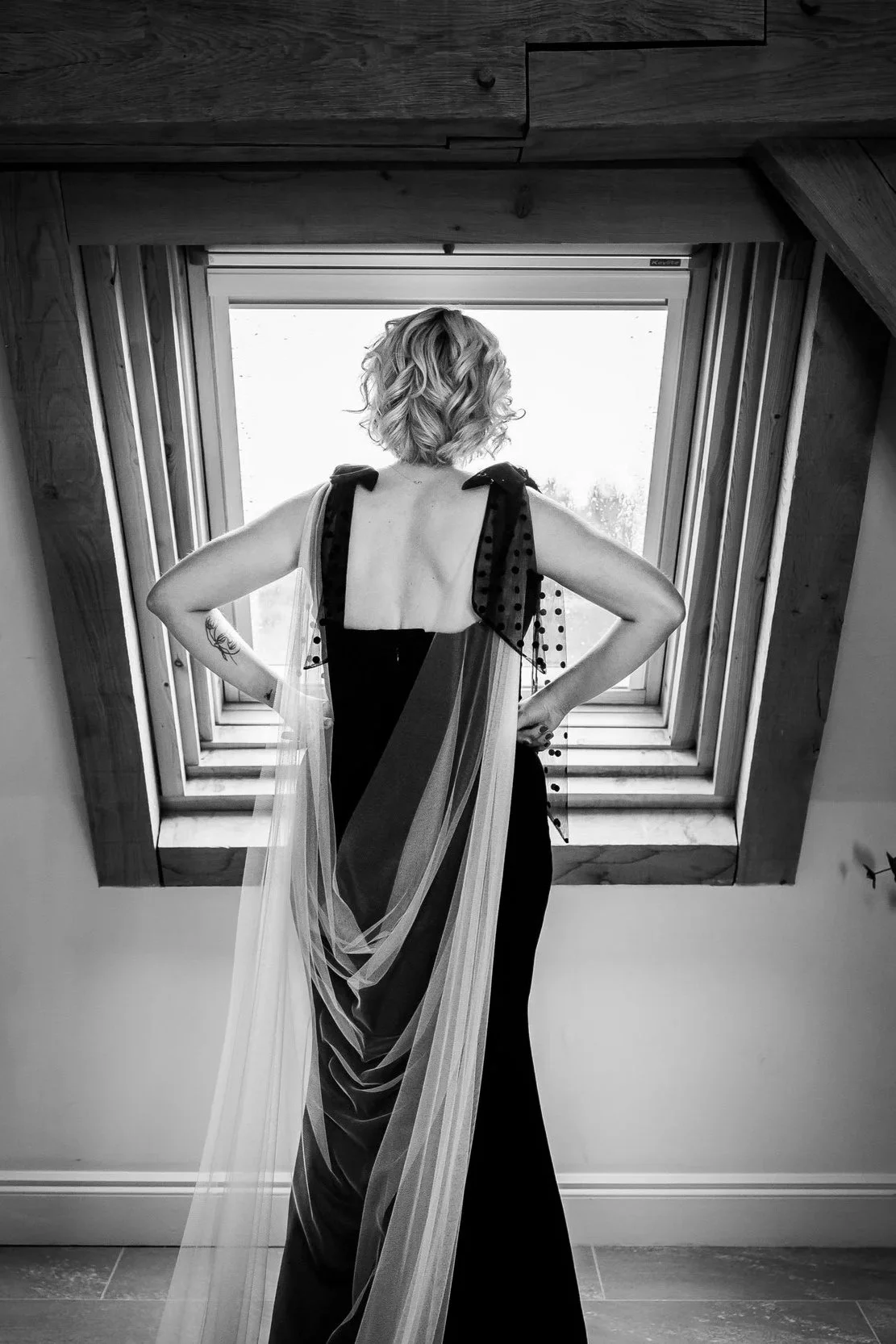 A woman in a gown standing in front of an attic window, looking outside, with wooden beams around her. Black and white photo.