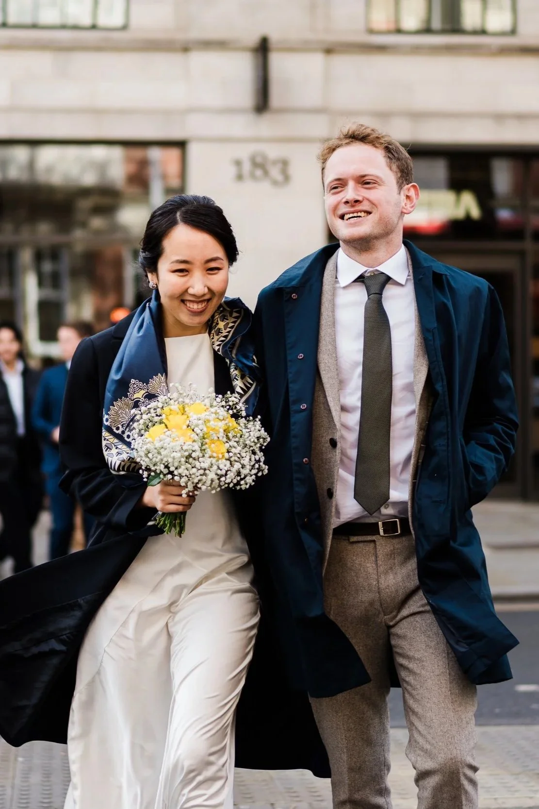 A couple walking outdoors, smiling, with the woman holding a bouquet of white and yellow flowers.