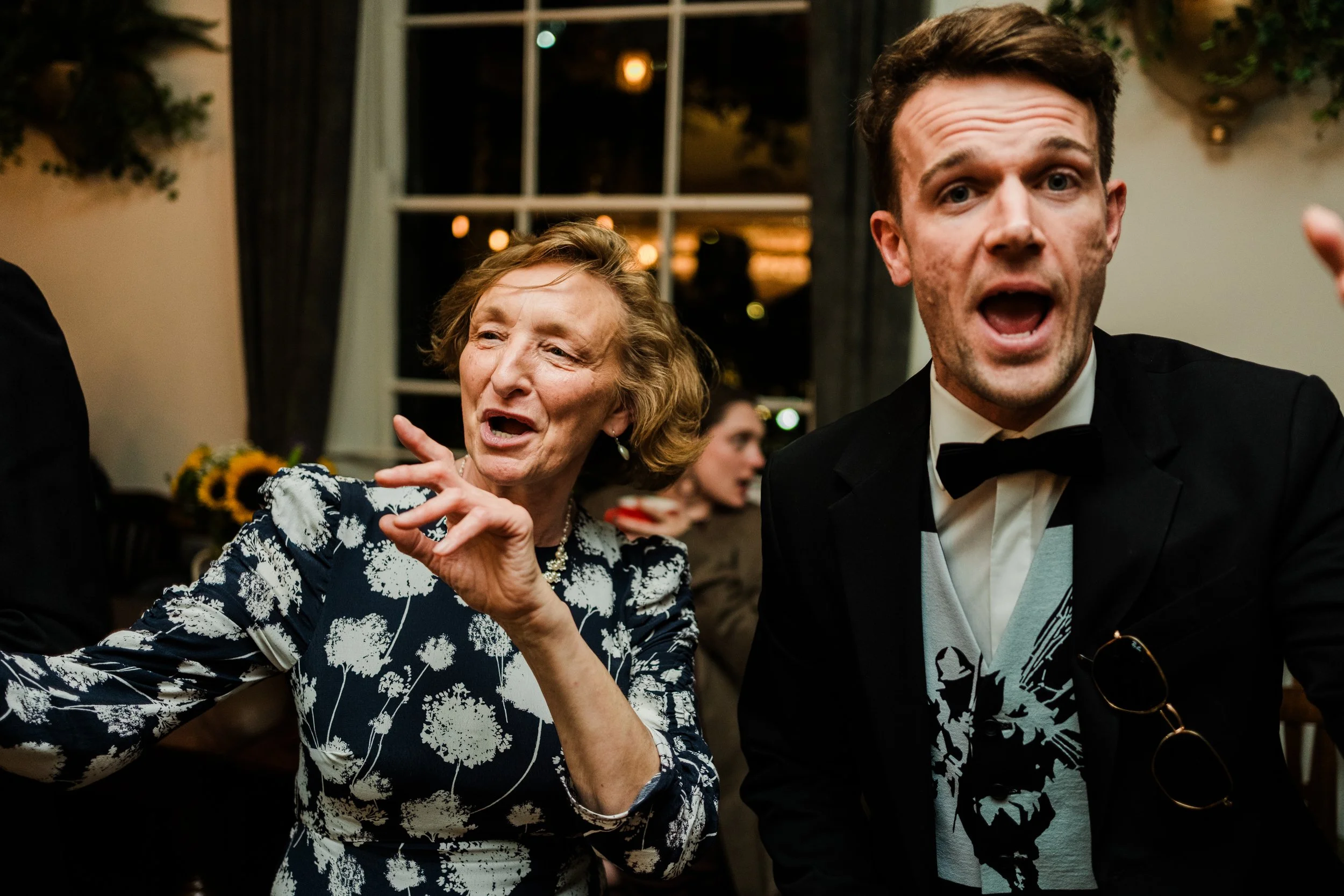 An older woman and a young man dressed in formal attire, engaging in a lively conversation or celebration at an indoor event, with the woman gesturing with her hand and the man wearing a tuxedo with a bow tie.