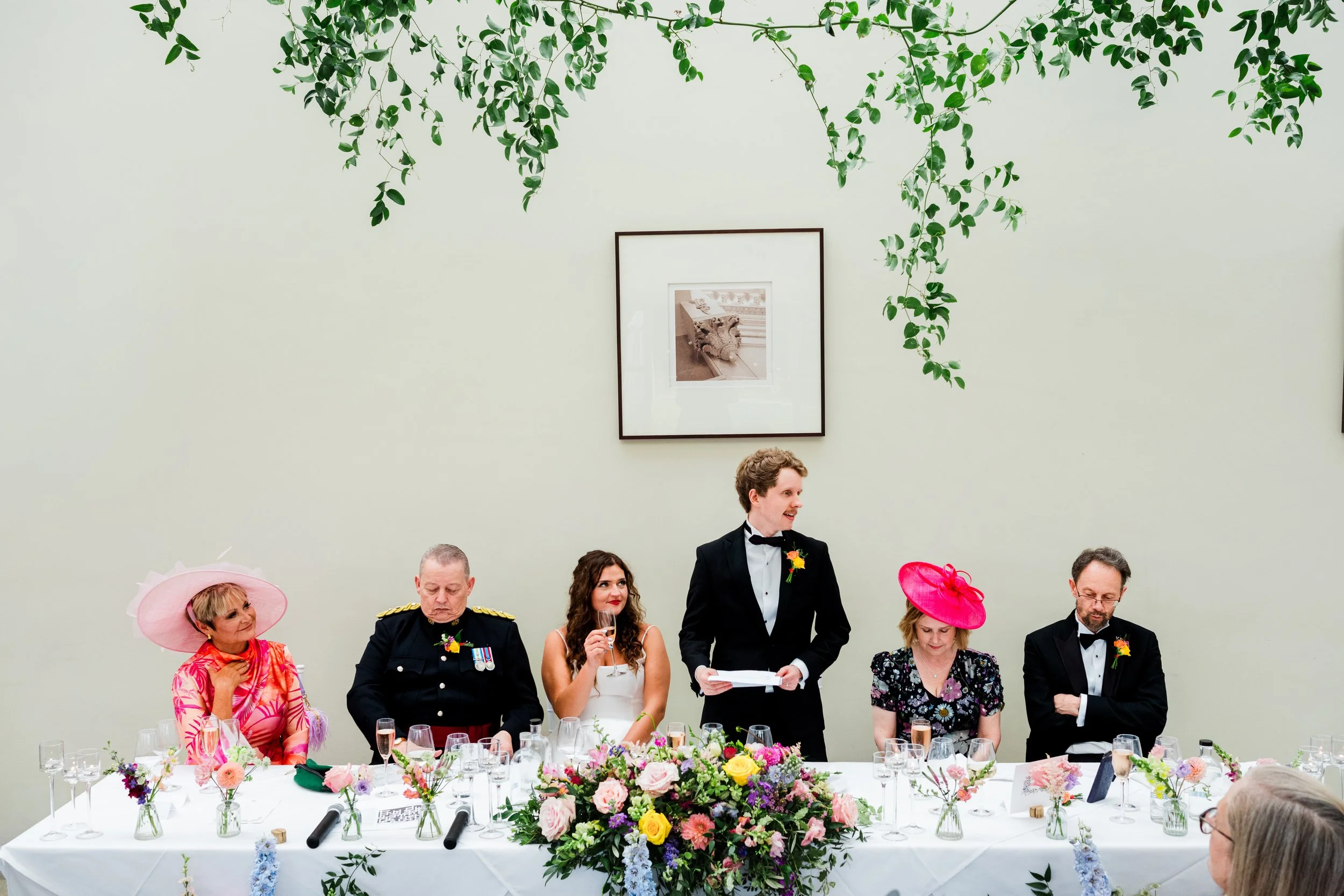People sitting at a long table with floral arrangements, dressed in formal attire, with a man standing and giving a speech, at a wedding reception.
