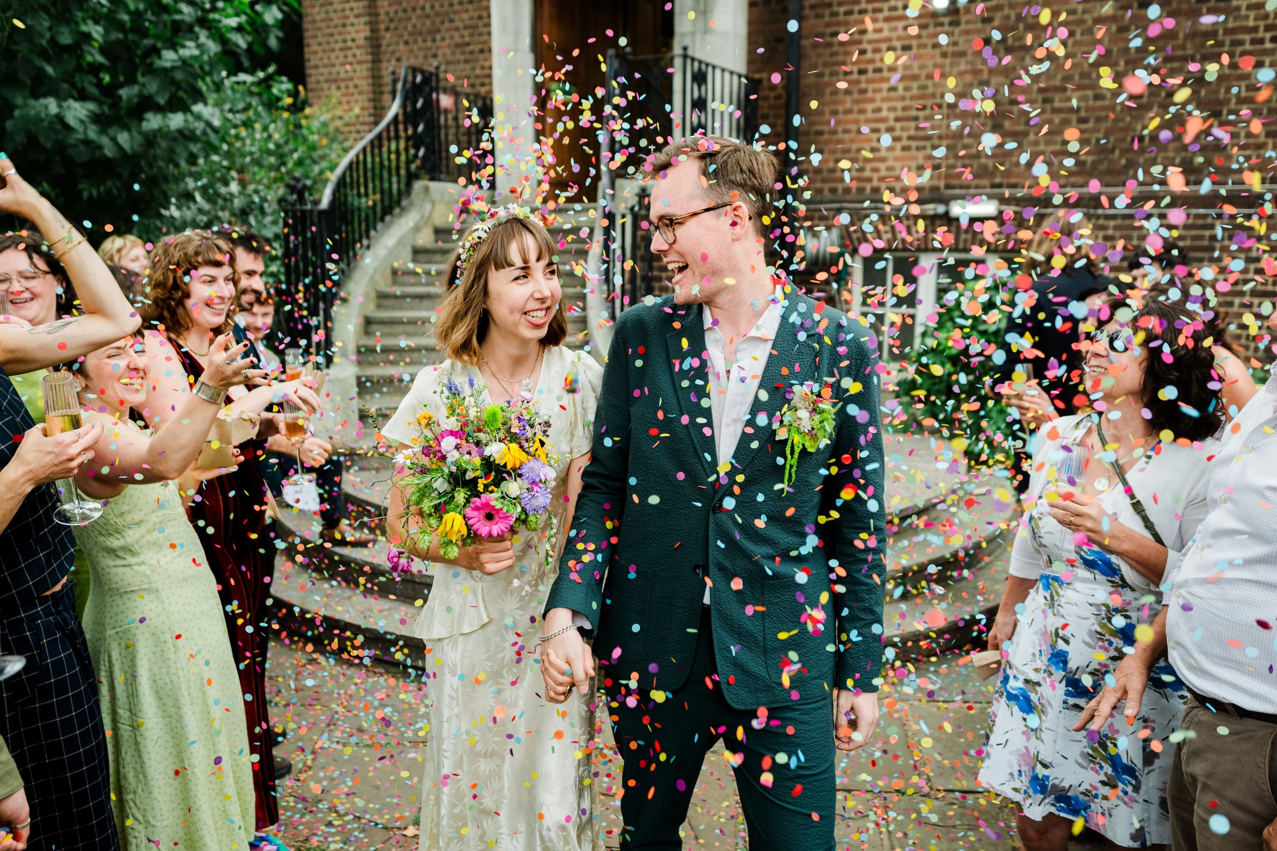 A newly married couple celebrates their wedding outdoors, surrounded by friends throwing colorful confetti. The bride holds a bouquet of flowers, and the groom is dressed in a dark suit. Guests are smiling and taking photos, creating a joyful atmosph