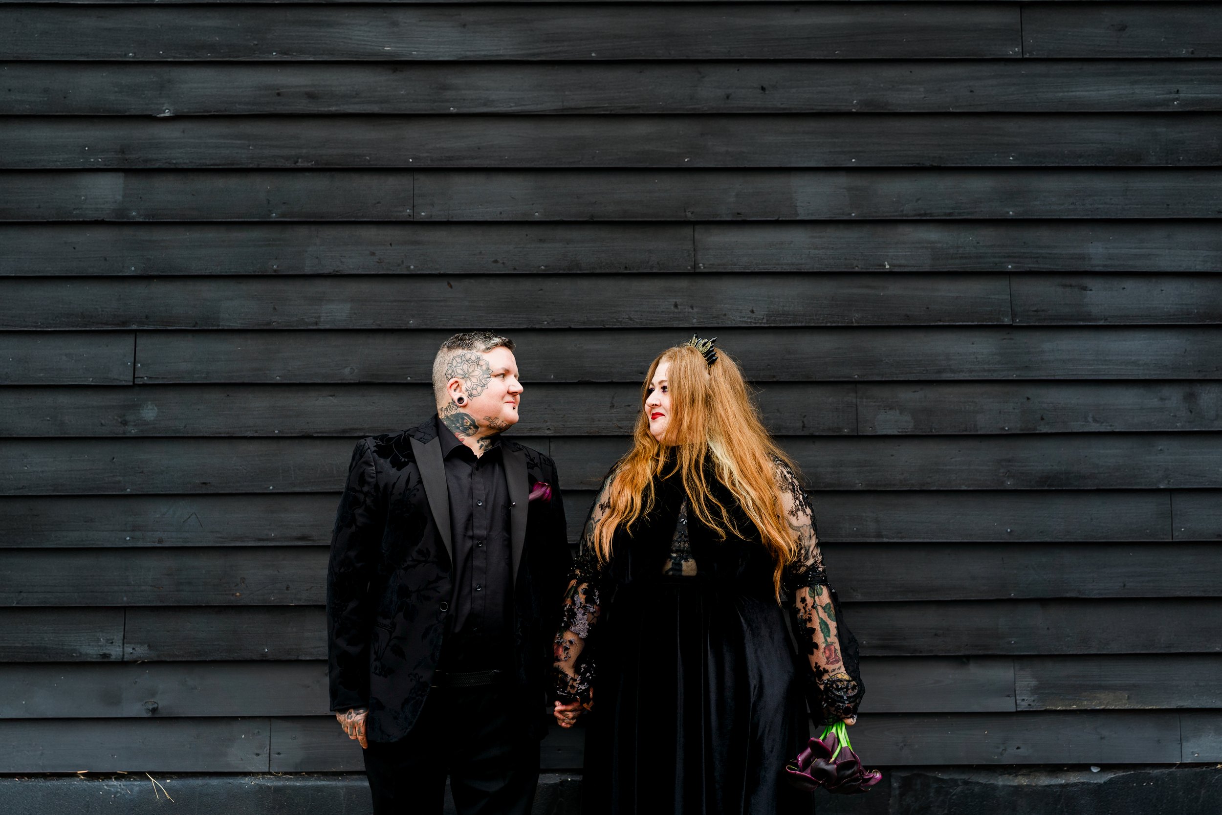 A couple with tattoos dressed in black clothing holds hands in front of a black wooden wall, looking at each other.