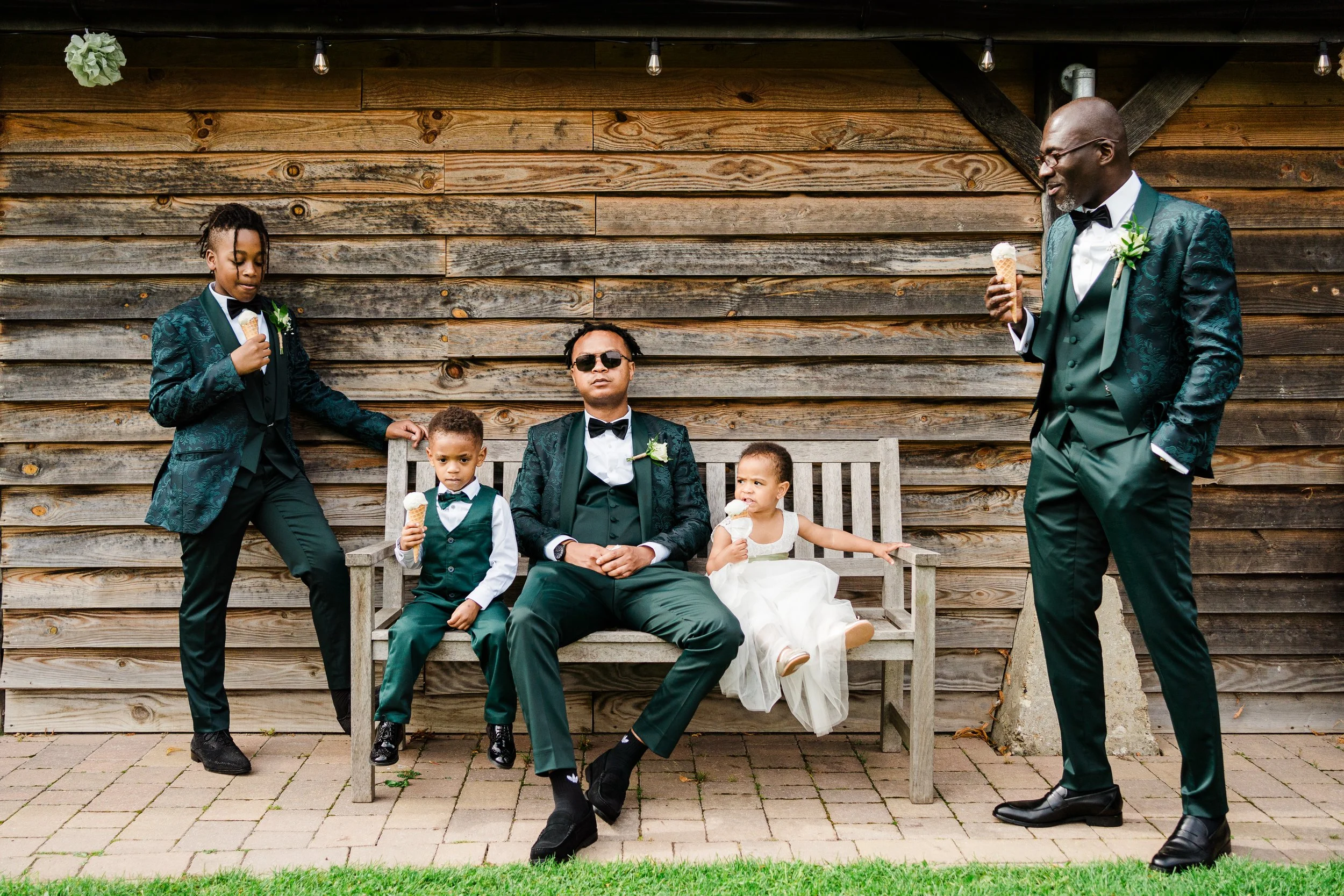 A group of six people, including three children and two adult men, dressed in formal suits and dresses, sitting and standing on a wooden bench and ground outdoors against a wooden wall. They are enjoying ice cream cones.