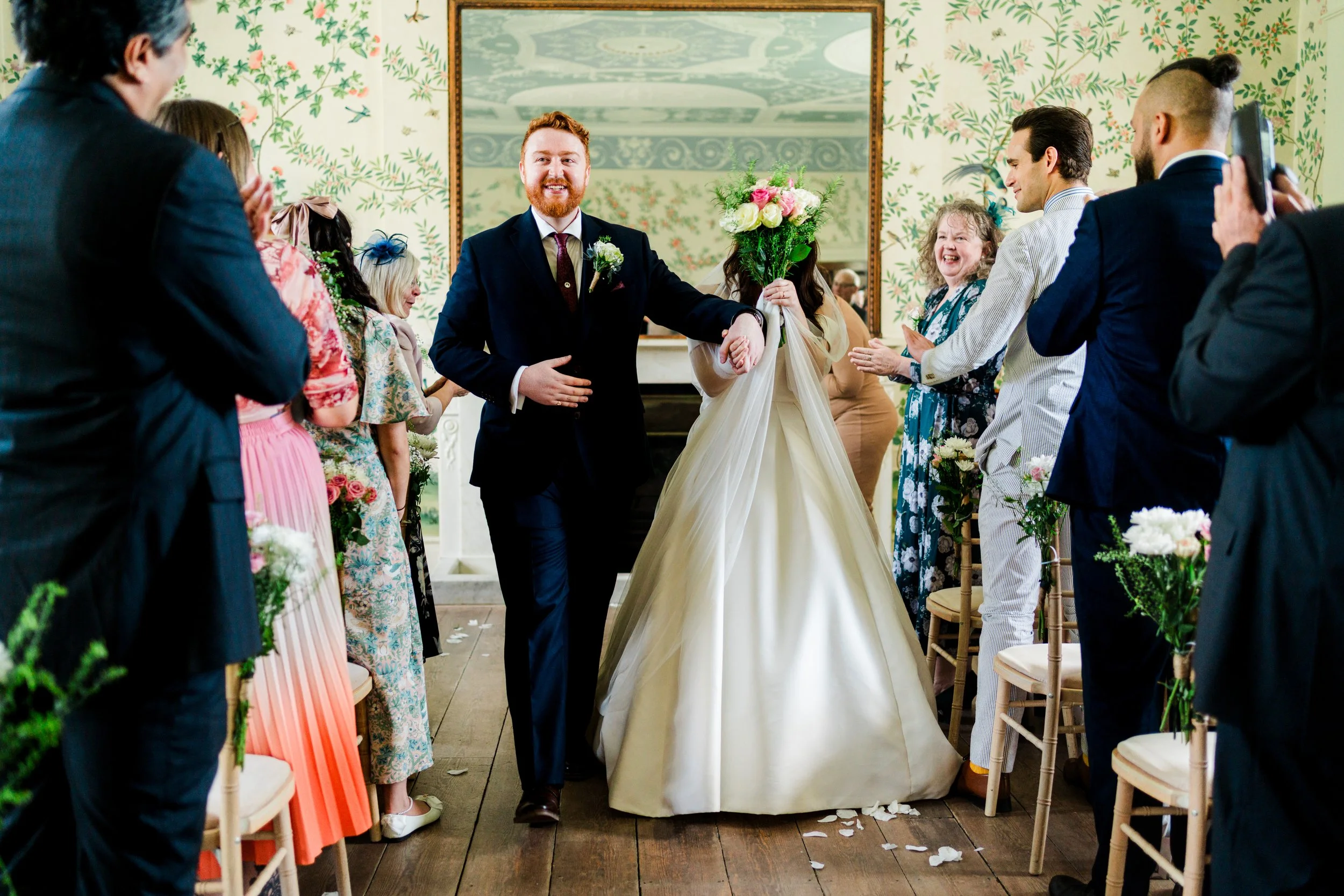 A wedding ceremony with a bride in a cream dress holding a bouquet and a groom in a dark suit, walking down the aisle together, surrounded by guests in colorful attire applauding. The setting is decorated with floral arrangements and a large mirror o