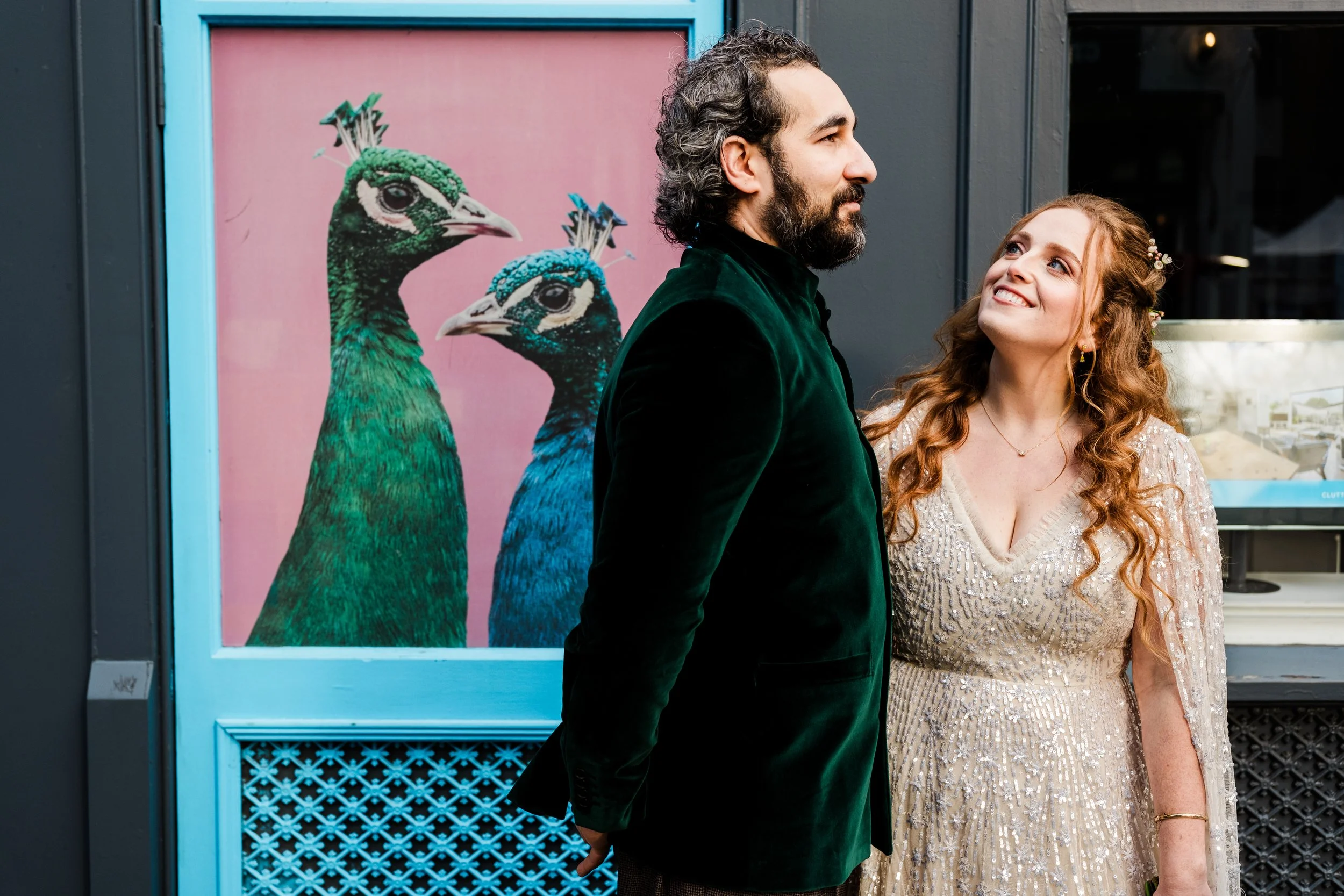 A man and woman stand close to each other outside, with a colorful peacock artwork in the background.