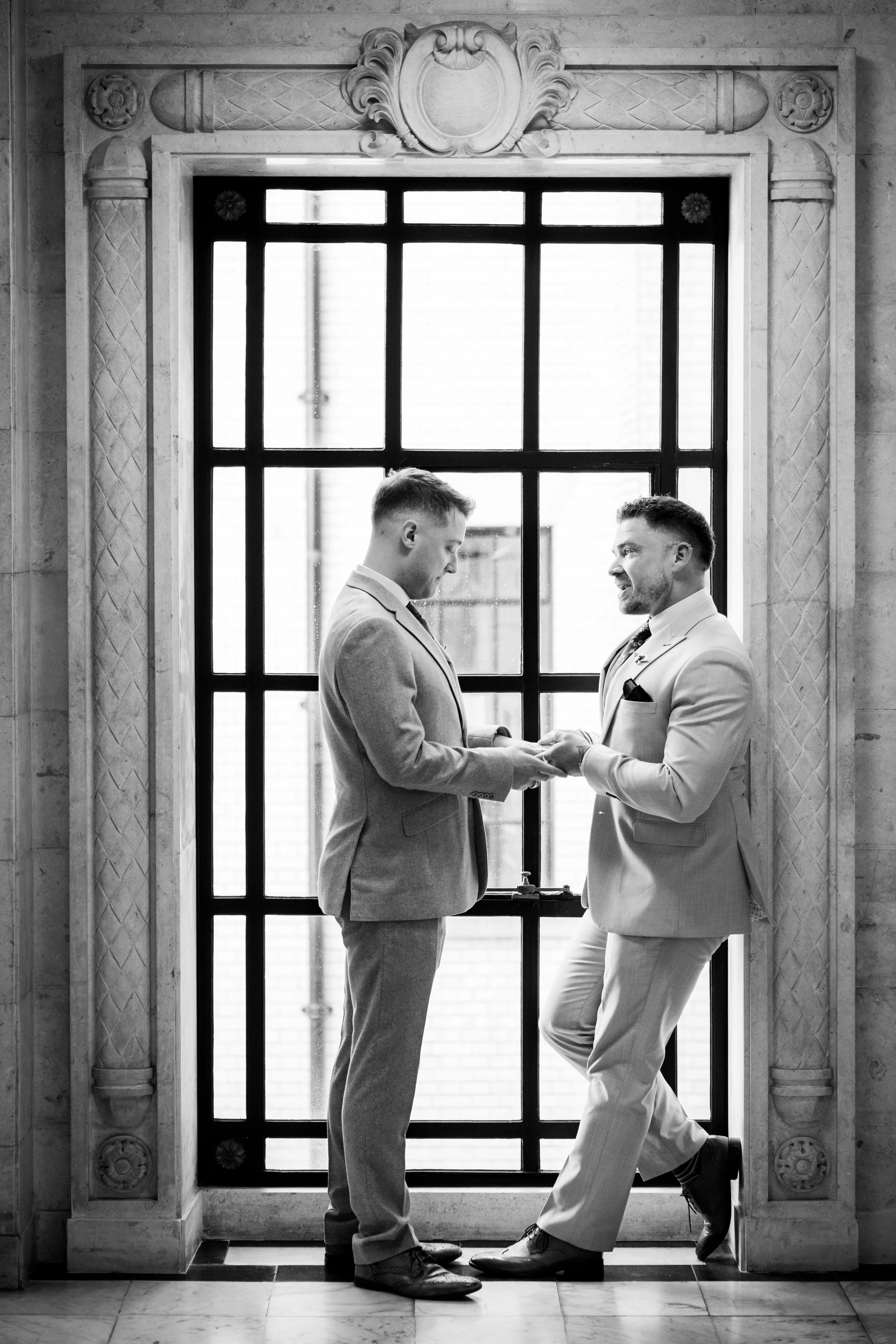 Two men in suits share a moment, holding hands, inside a building with a large window behind them and ornate stone framing.