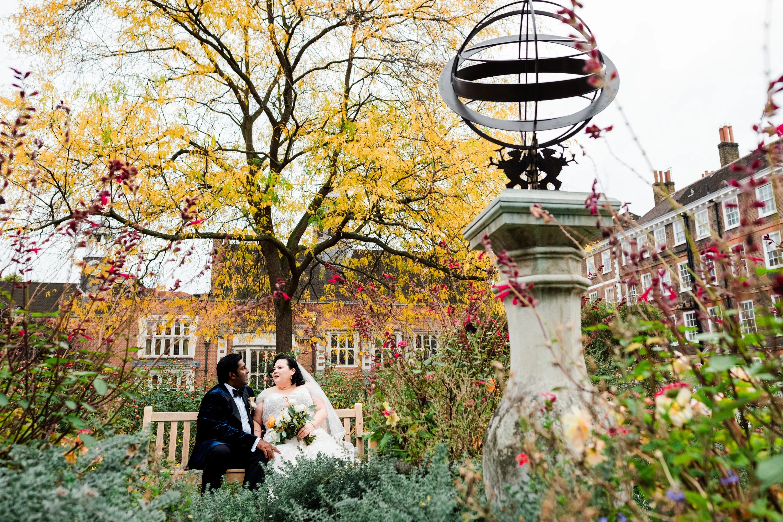 A bride and groom sitting on a wooden bench in a garden surrounded by autumn foliage, with a large yellow tree, colorful bushes, and a decorative black metal globe sculpture on a stone pedestal.