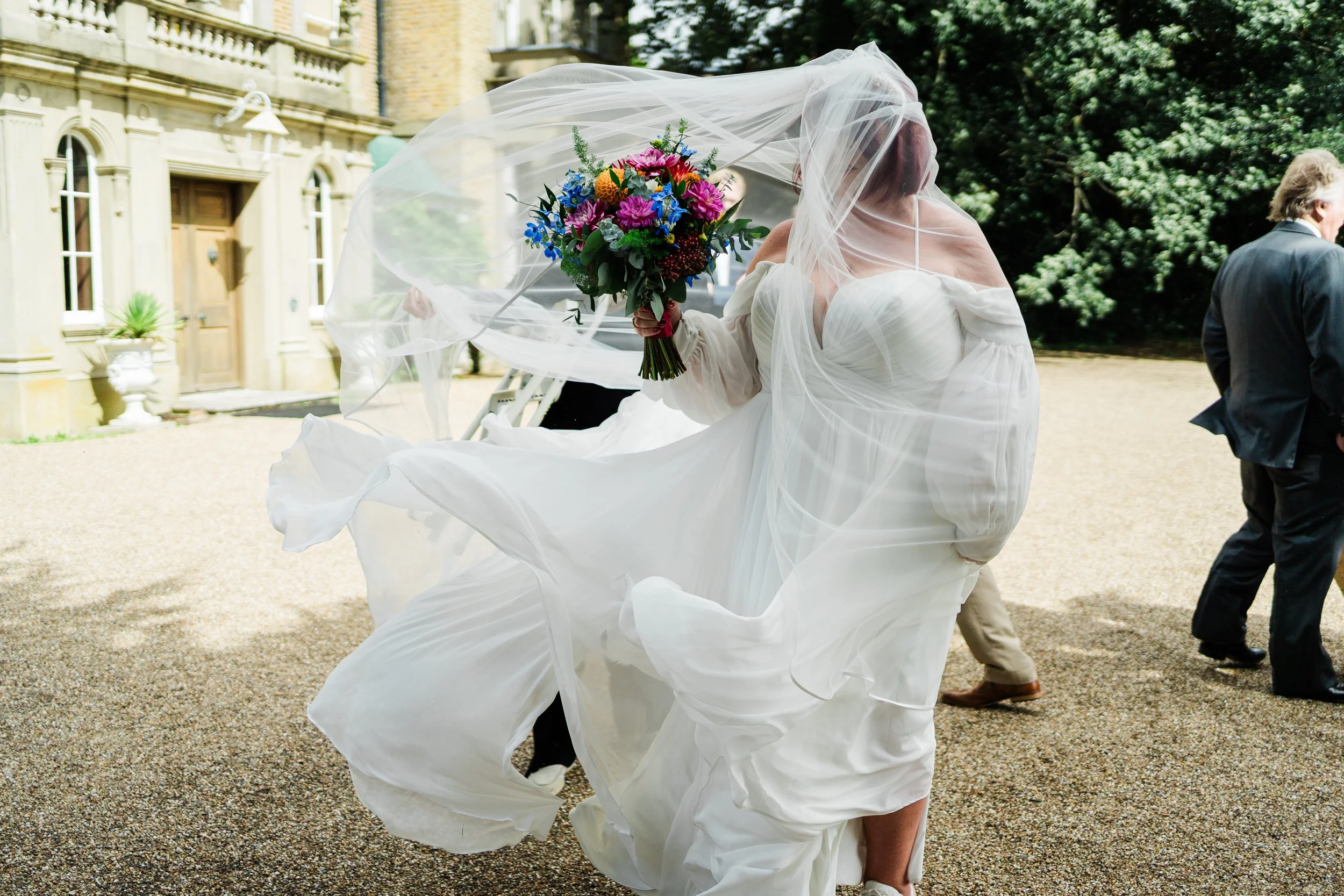 Bride in a white wedding dress with a veil holding a colorful bouquet of flowers, outdoors during a wedding celebration.