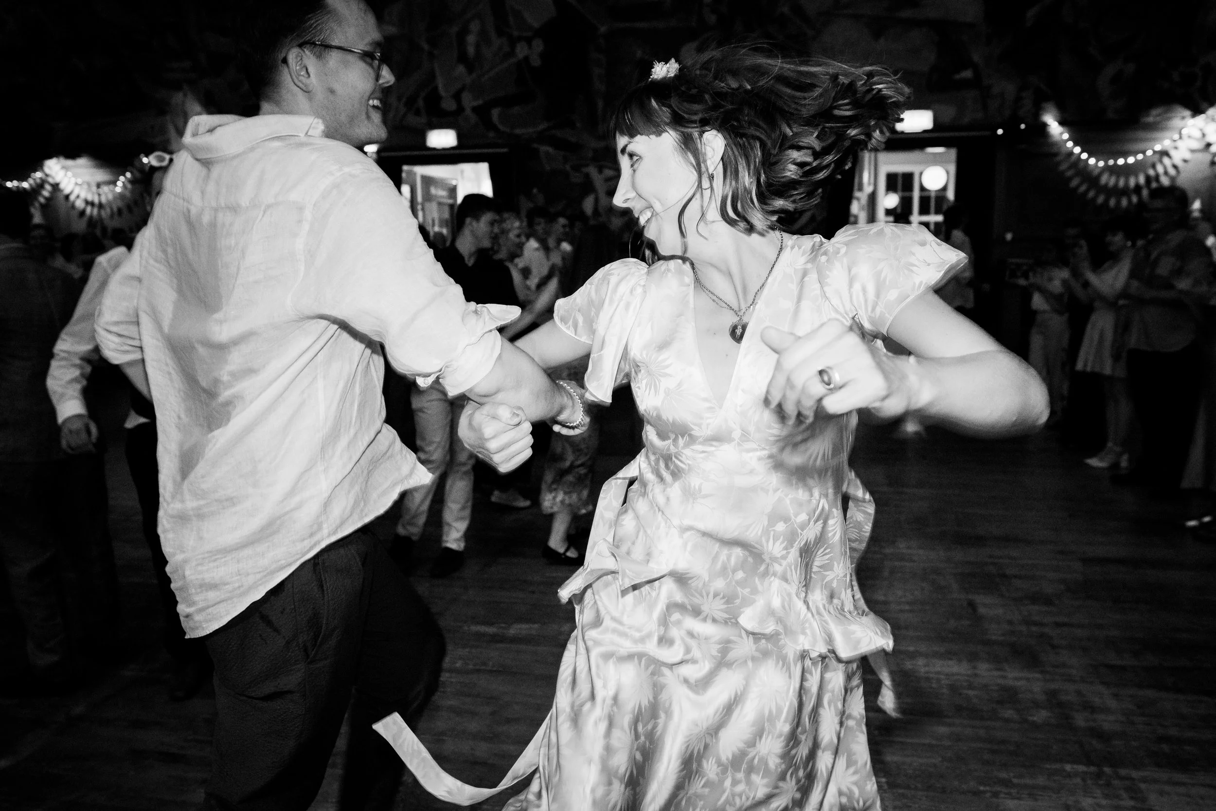 Two people dancing, a man and woman, in a lively indoor setting with other people in the background. The woman has long hair flowing as she spins, and they are holding hands.