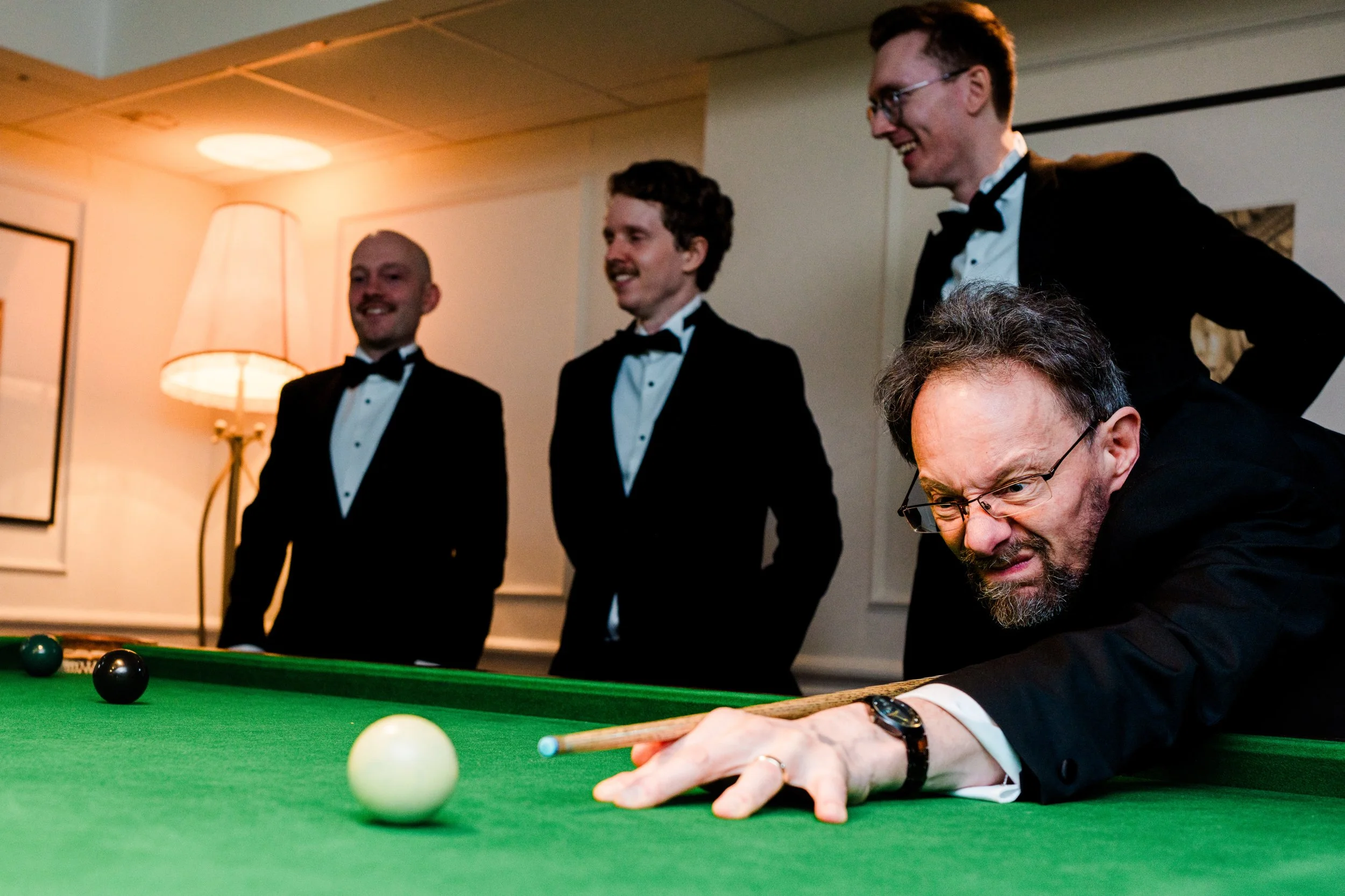 A man wearing glasses and a black suit plays pool, concentrating as he aims to hit the cue ball. Four other men dressed in formal tuxedos stand in the background, watching and smiling in a room with warm lighting and a lamp.