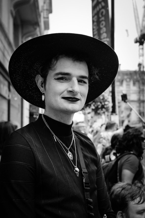 A woman with short hair, wearing dark lipstick, a large black sun hat, layered necklaces, earrings, and a black outfit, standing on a busy city street in black and white.