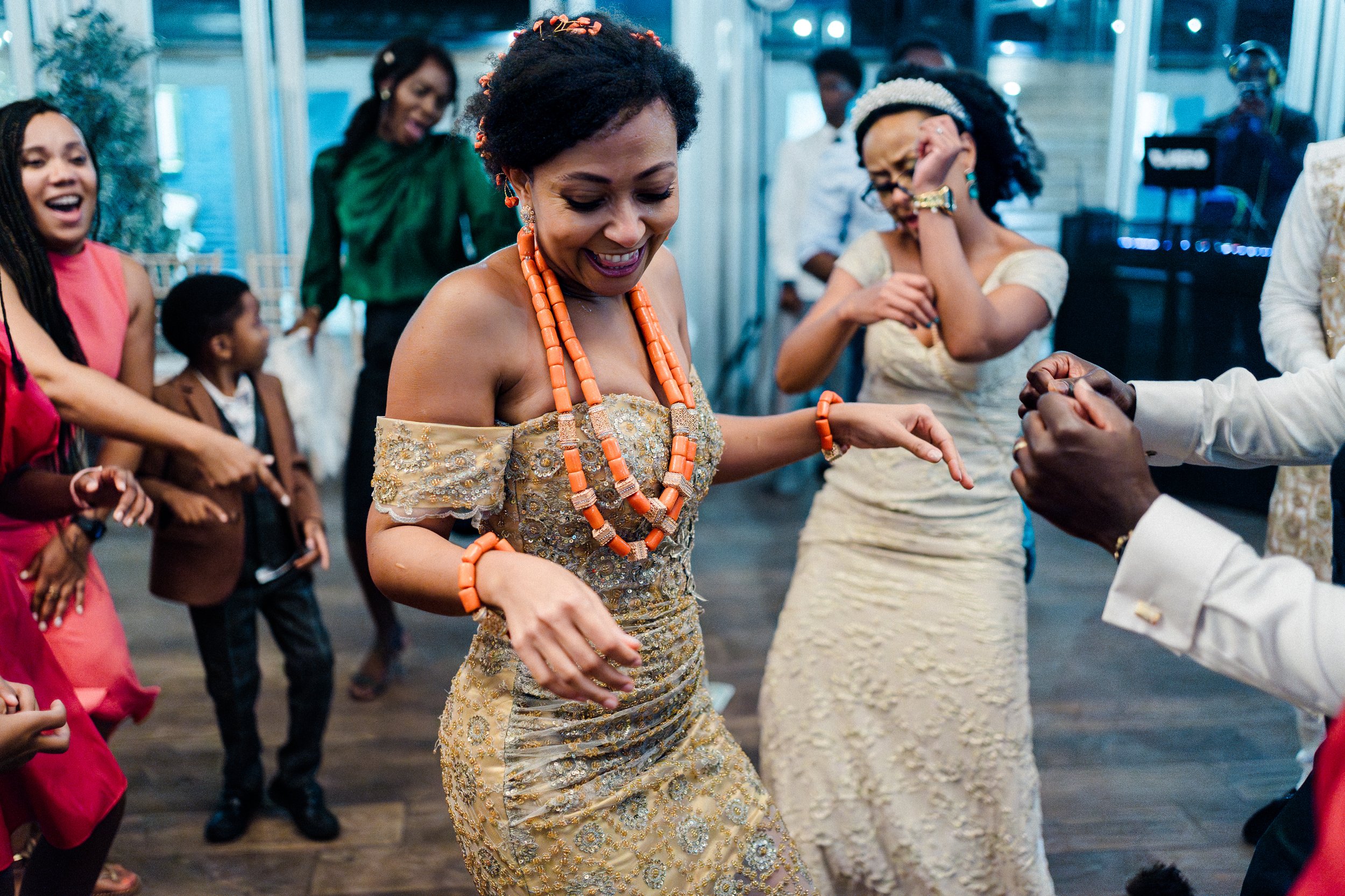 People dancing at a celebration, with black women wearing elegant dresses and colorful jewelry, smiling and enjoying themselves indoors.