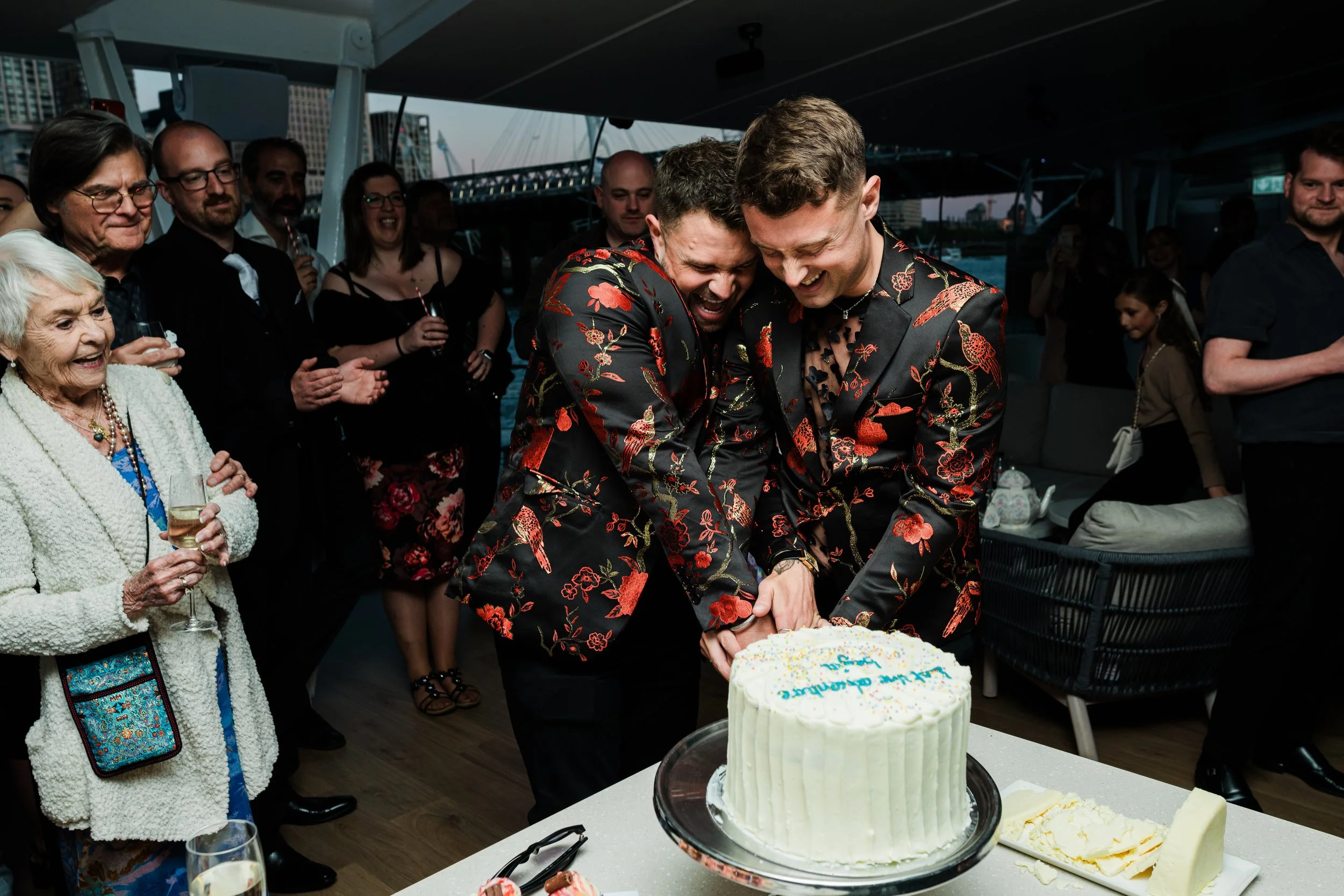 Two men in matching floral suits cutting a cake at a wedding reception, surrounded by smiling guests on a boat.