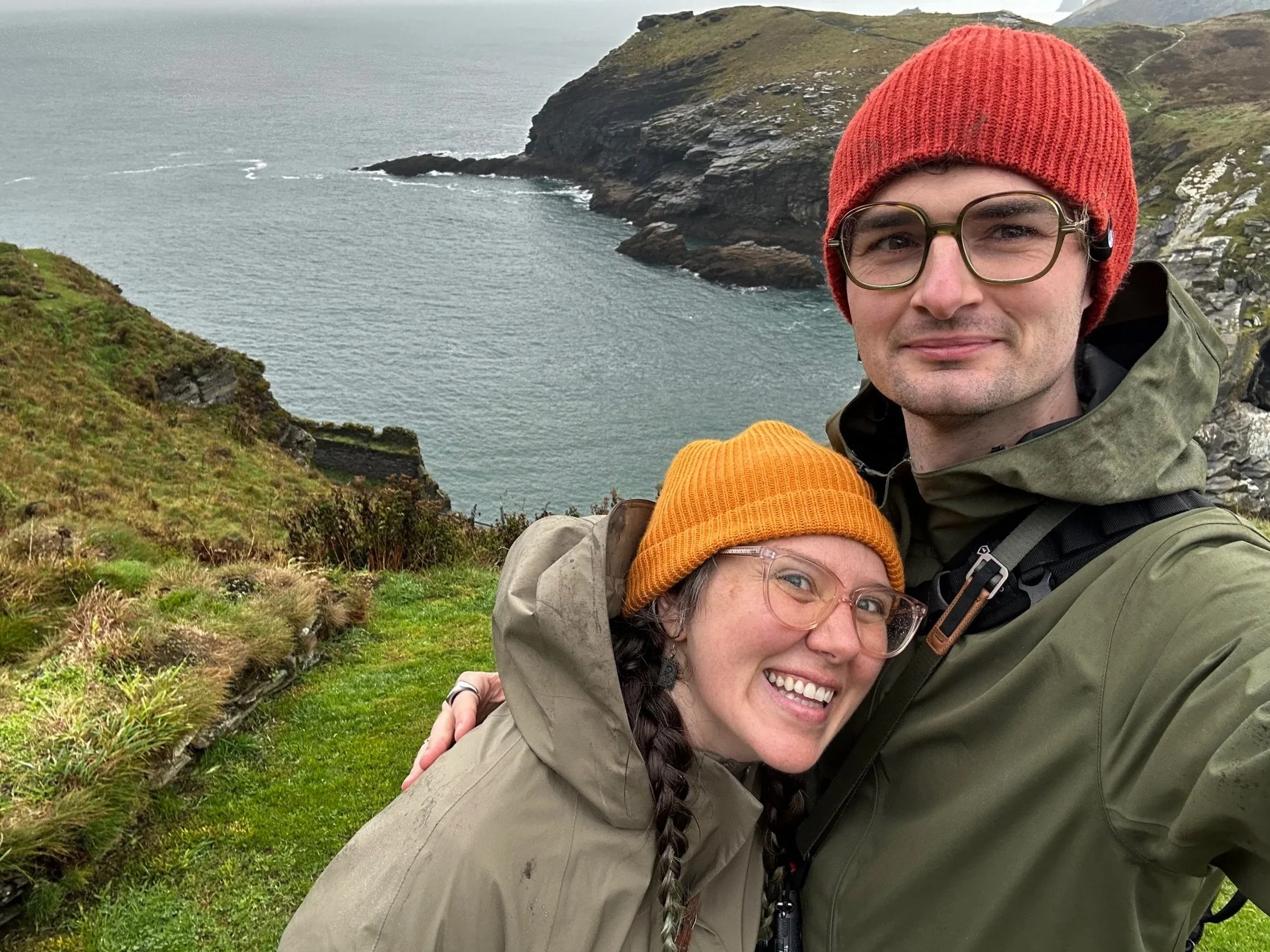 A smiling couple wearing outdoor jackets and knit hats takes a selfie on a grassy hillside overlooking a river or fjord with rocky cliffs.