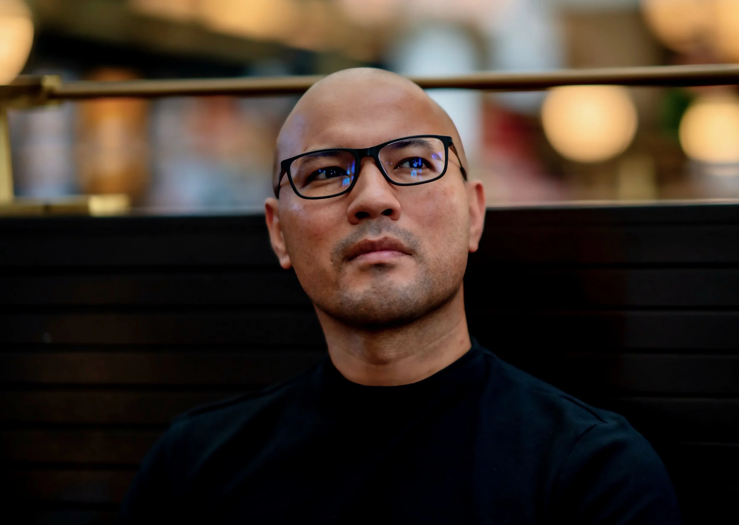 A bald man wearing glasses and a black shirt, sitting in front of a black wooden background with blurry lights in the distance.