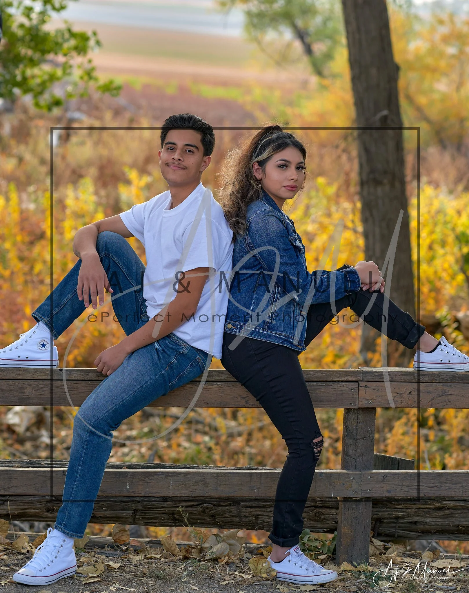 A young man and woman sit back-to-back on a wooden railing outdoors during autumn, with fall-colored trees in the background, both wearing casual clothing and white sneakers.