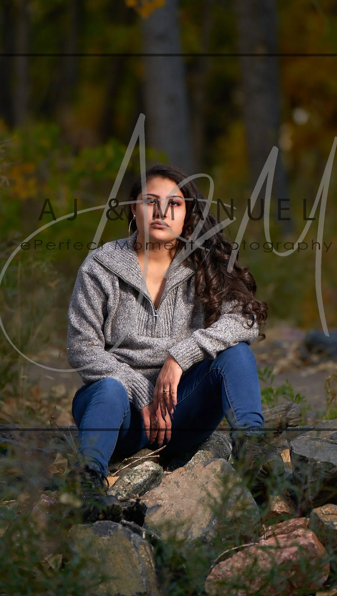 A woman with long, curly dark hair is sitting outdoors on rocks in a forested area during autumn. She is wearing a gray sweater and blue jeans, looking thoughtfully at the camera.