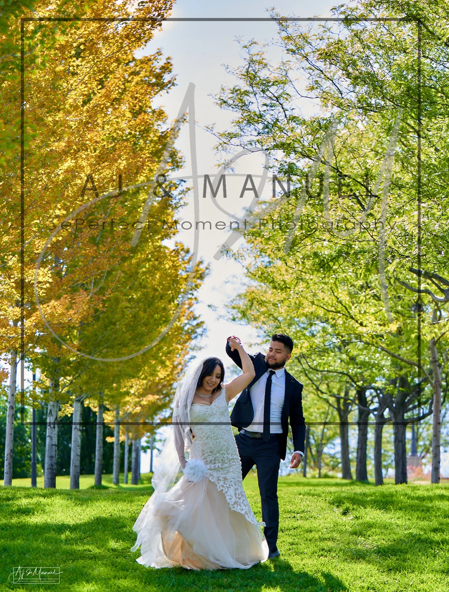 A bride and groom dancing outdoors on a grassy field surrounded by green and yellow tree leaves on a sunny day.