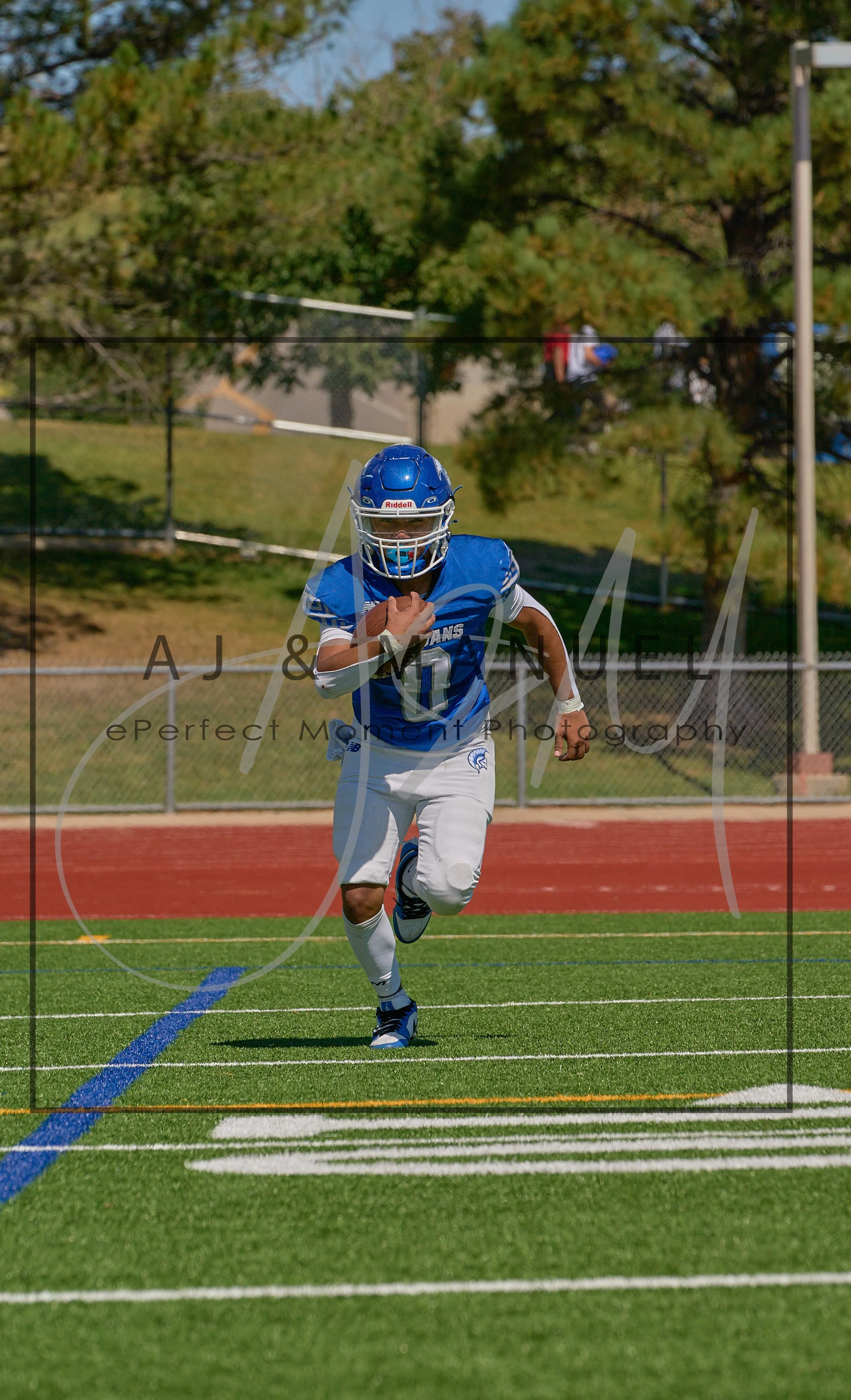 A young football player wearing a blue helmet, jersey, and white pants is running on a football field with a football in his hand. The background shows a track, chain-link fence, orange and green trees, and a clear blue sky.