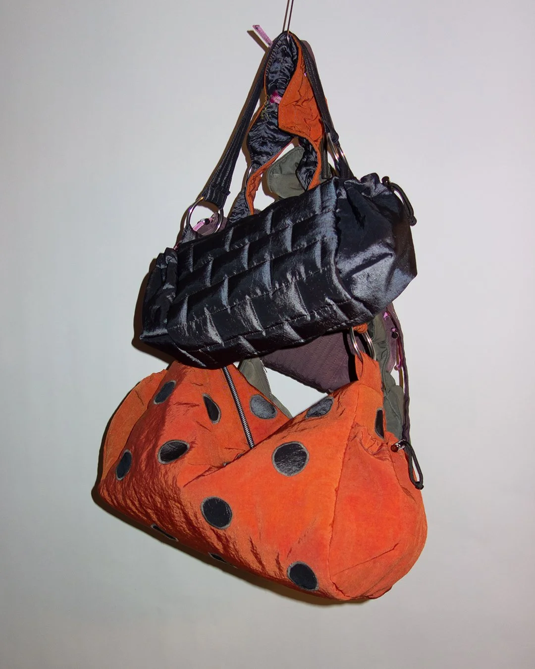Suspended collection of various bags including a black quilted bag, an orange waterfowl-shaped bag with black polka dots, and a small gray and orange pack, hanging from hooks against a plain background.