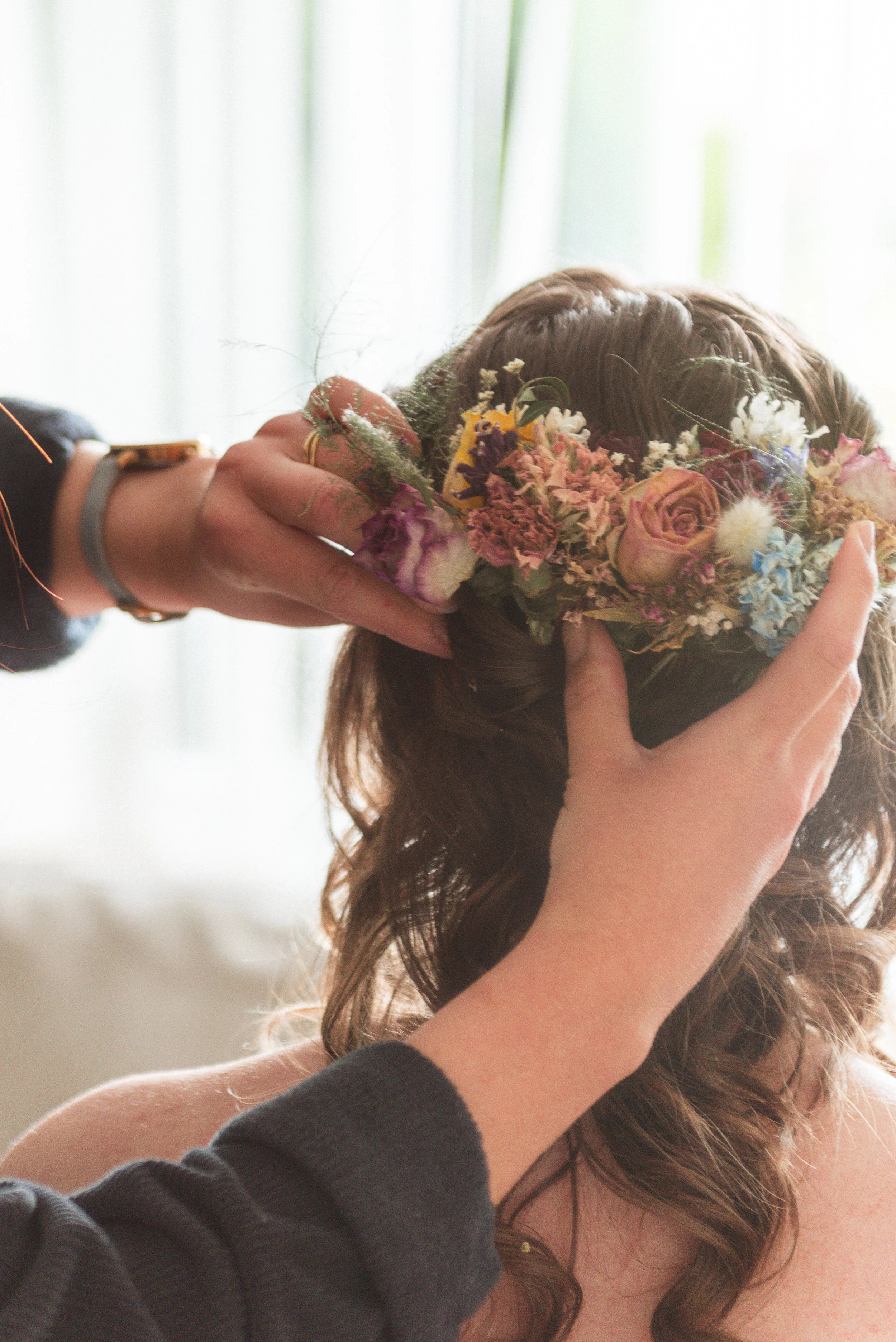 a hairdresser adjusts florals in a bridal up do with soft morning light streaming in