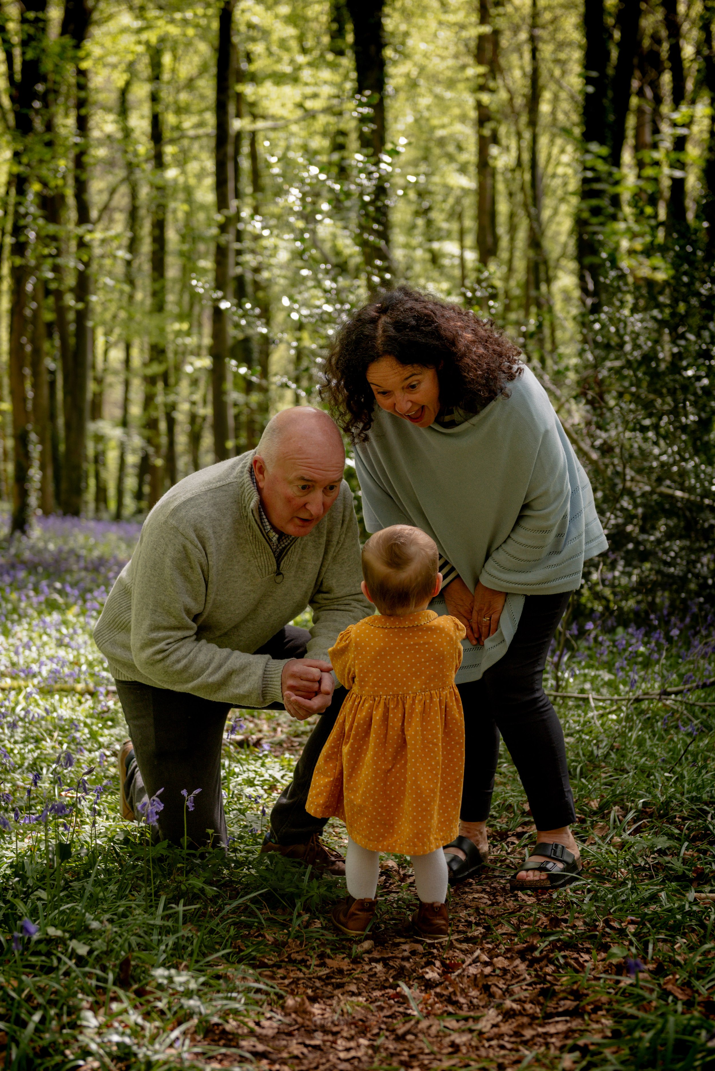 Grandparents crouch down to talk to their toddler granddaughter in a bluebell carpeted woodland clearing
