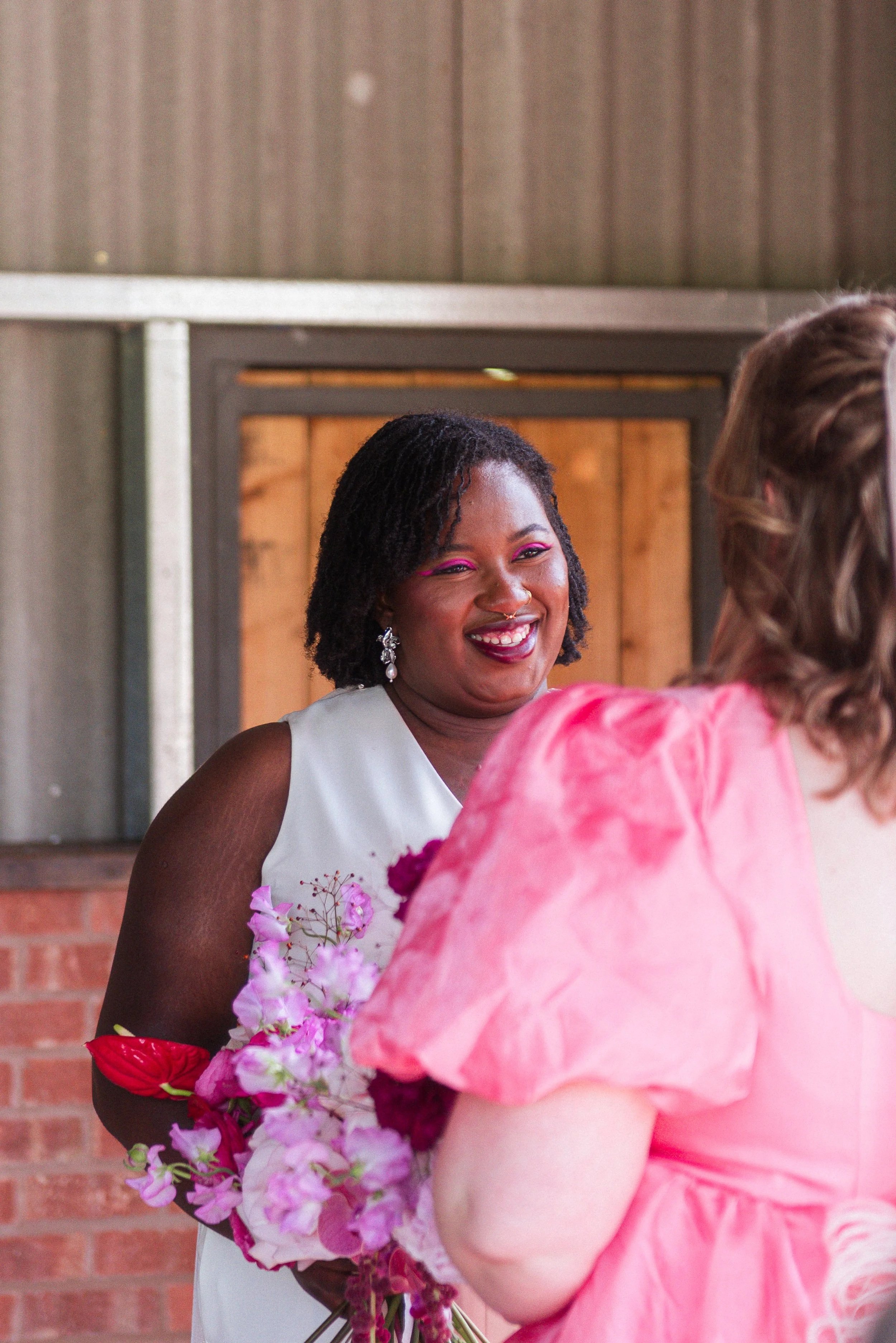 A woman stands facing her bride to be, who is holding a bright pink bouquet and wearing a bright pink non-traditional wedding dress