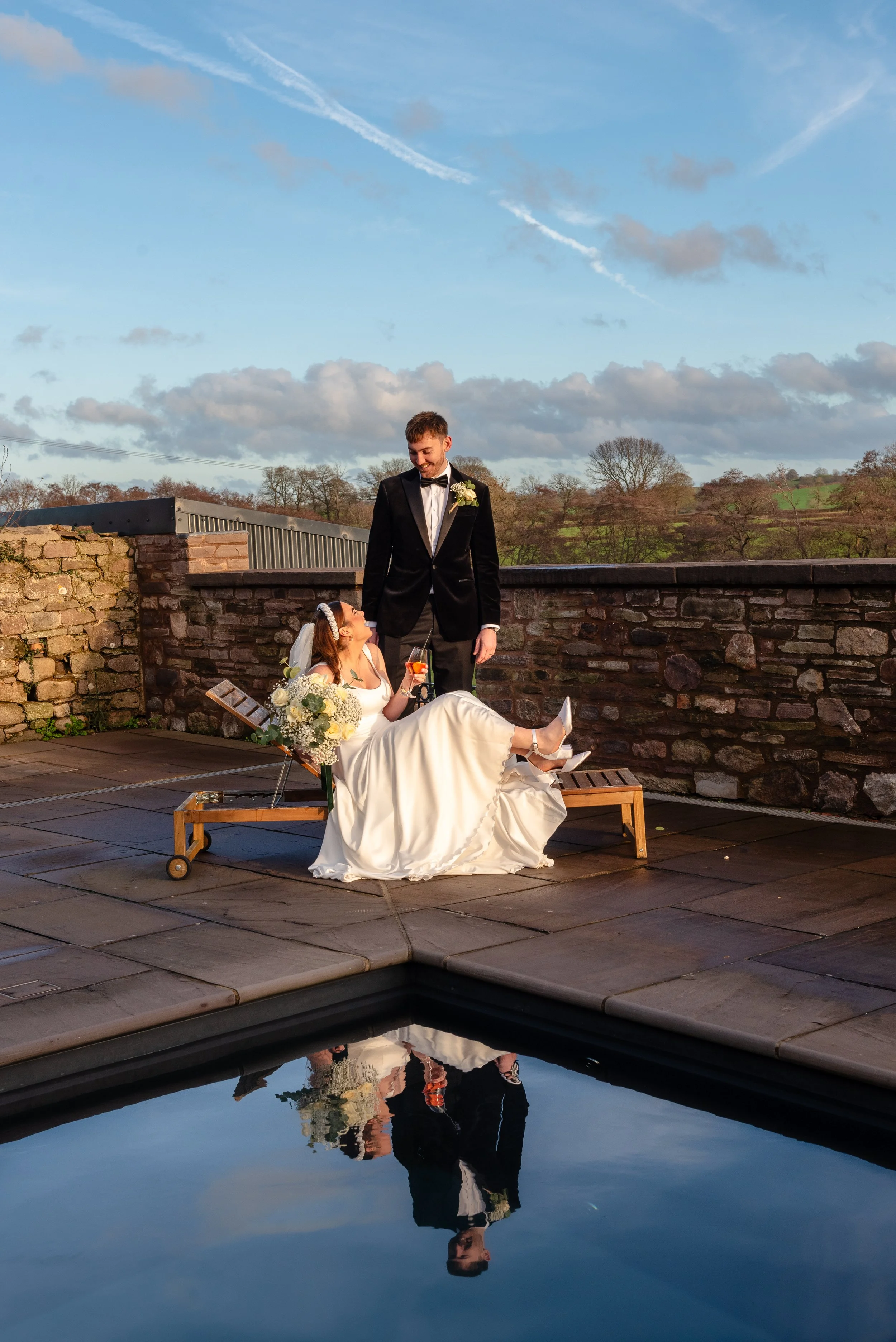 A stylish couple of newly weds pose with a cocktail in front of the pool laughing at each other