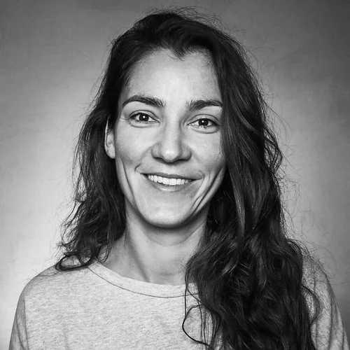 Sort-hvid portrætfotografi af en smilende ung kvinde med langt, krøllet hår, iført en casual grå T-shirt, mod en neutral baggrund.