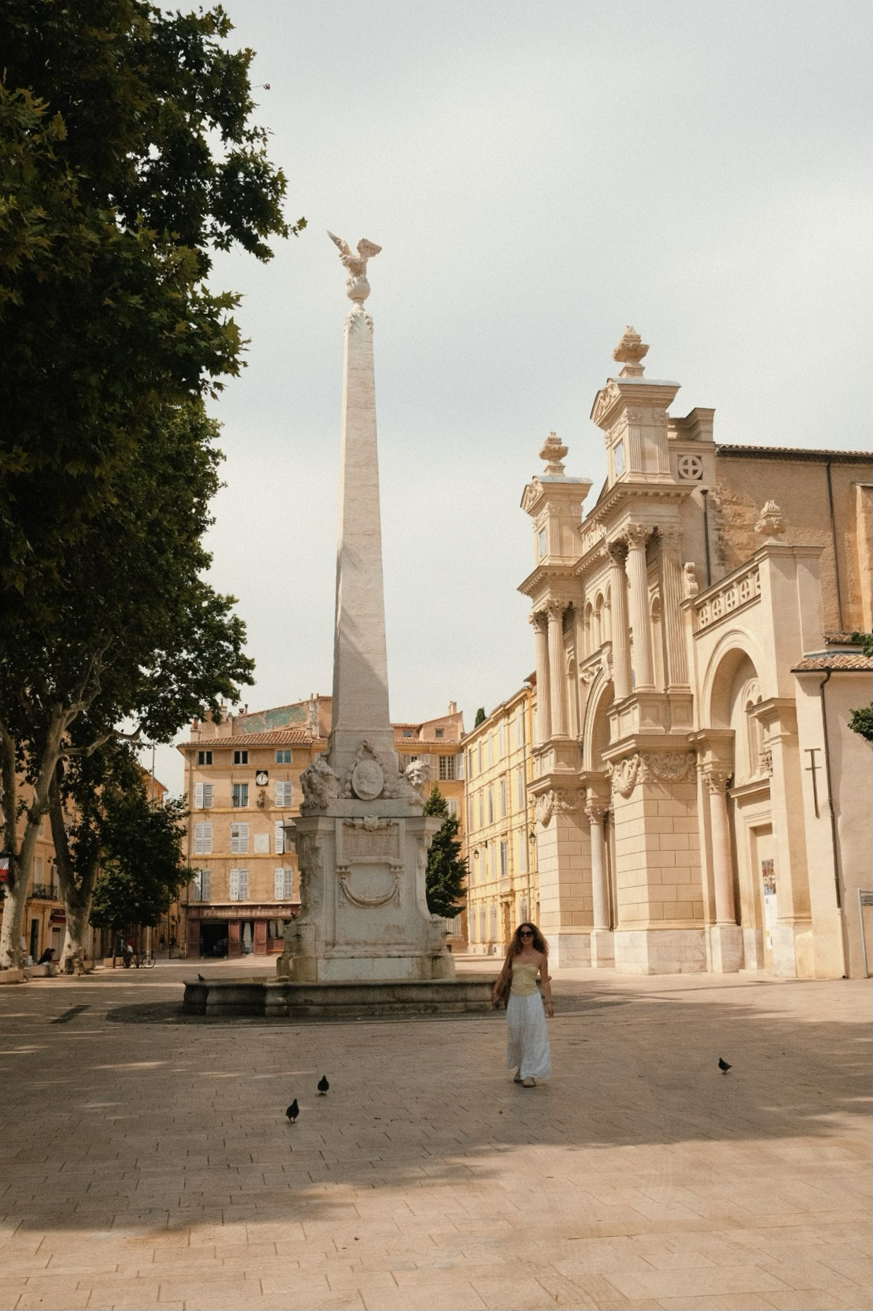 Place avec une colonne en marbre ornée de sculptures et un bâtiment baroque en arrière-plan. Une femme marche sur la place, entourée de quelques pigeons, sous un ciel clair.