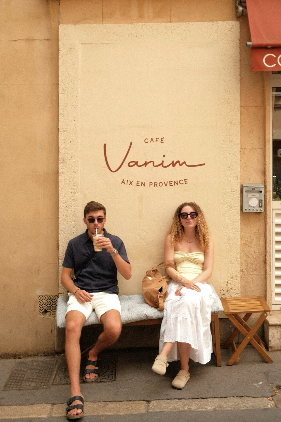 Deux personnes assises sur un banc devant un mur avec une enseigne de café. L'homme tient une boisson et porte des lunettes de soleil, la femme porte également des lunettes de soleil et une robe blanche. Café Vanim à Aix en Provence