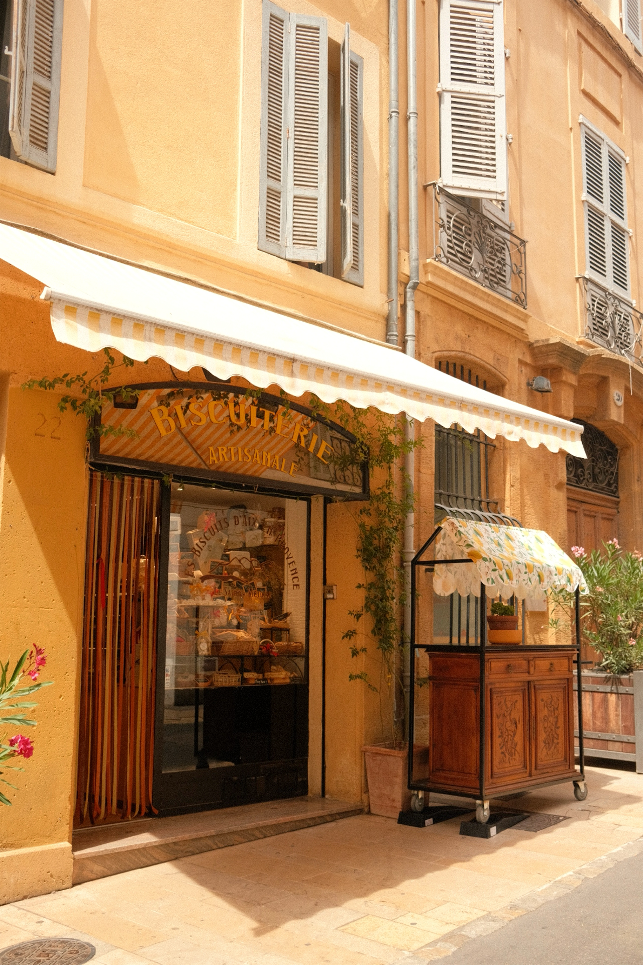 Façade d'une boulangerie artisanale avec une enseigne en arc, portant le mot 'Biscuiterie' en lettres jaunes. Un store rayé blanc et jaune est déployé au-dessus de l'entrée. Il y a une vitrine devant la boutique avec des produits de boulangerie, et u