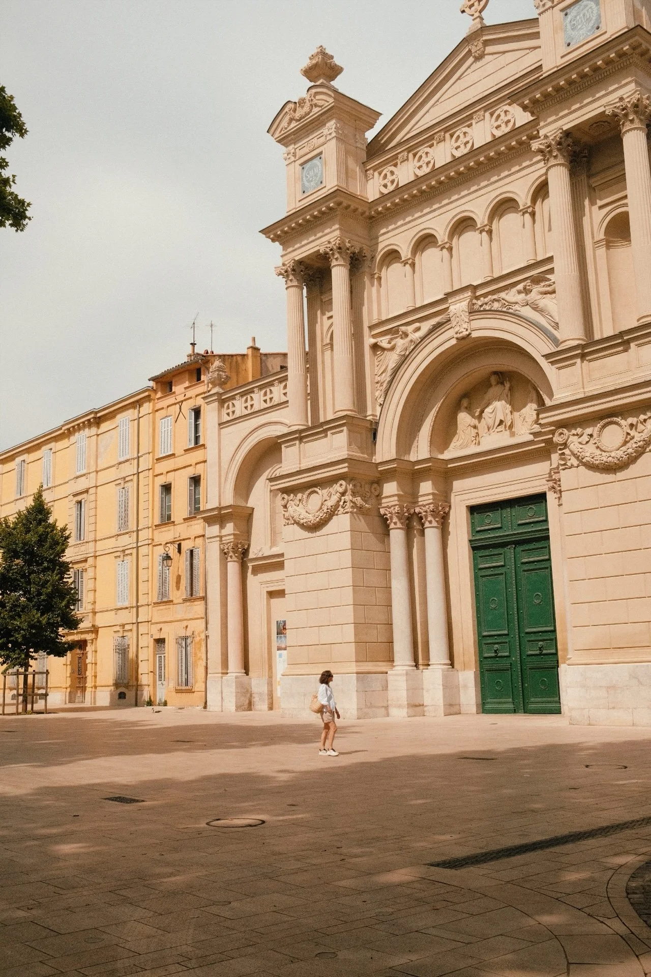 Une église ornée de sculptures et de colonnes, avec un grand portail vert, située dans une place pavée, avec une seule personne qui marche en bas.