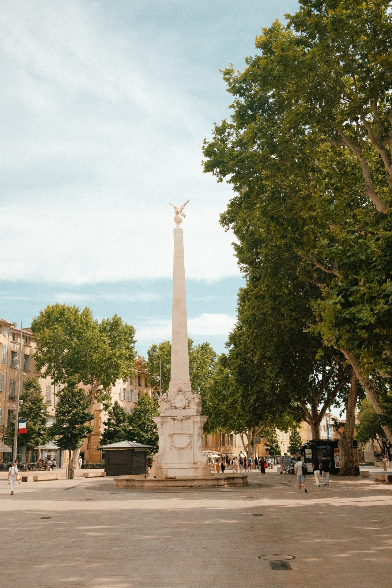 Une place pavée avec une colonne blanche ornée d'une sculpture d'un aigle en haut, entourée d'arbres verts et de bâtiments résidentiels, avec des personnes qui se promènent à Aix en Provence, Sud de la france