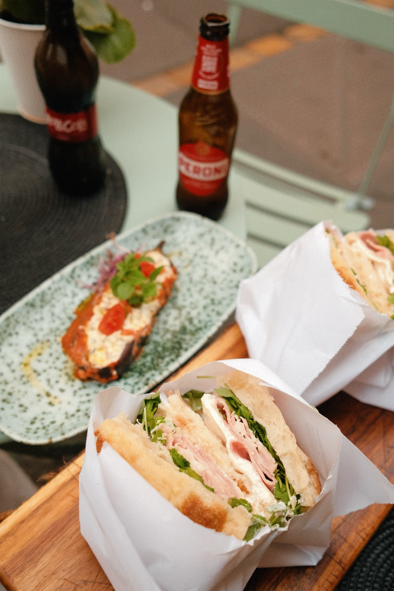 Sandwichs, une bouteille de bière, et une assiette avec une portion de roasted eggplant topped avec de la sauce, du fromage, et une garniture verte, sur une table en bois.
