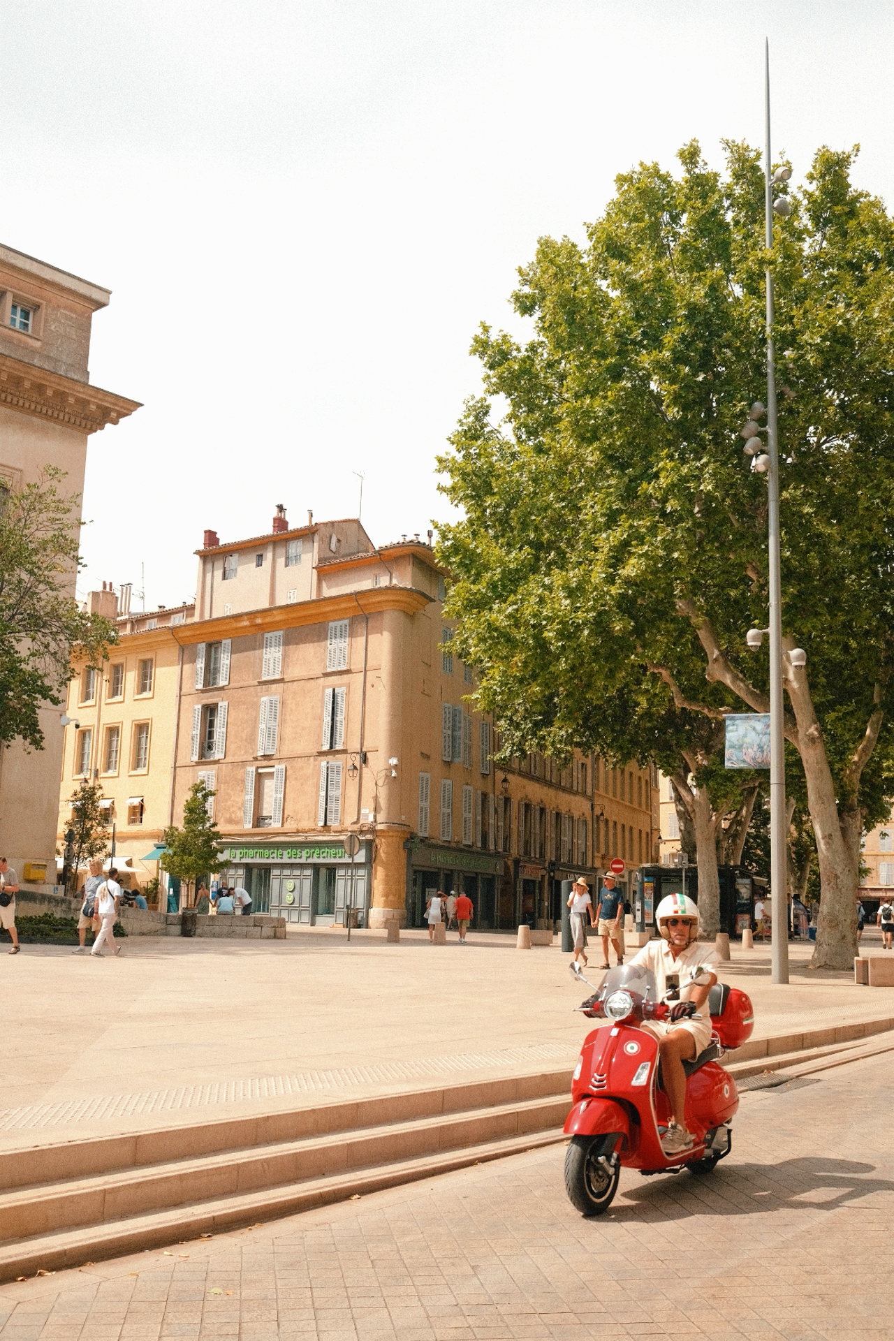 Une place urbaine ensoleillée avec des immeubles en arrière-plan, des arbres, des piétons et une personne roulant en scooter rouge portant un casque blanc.