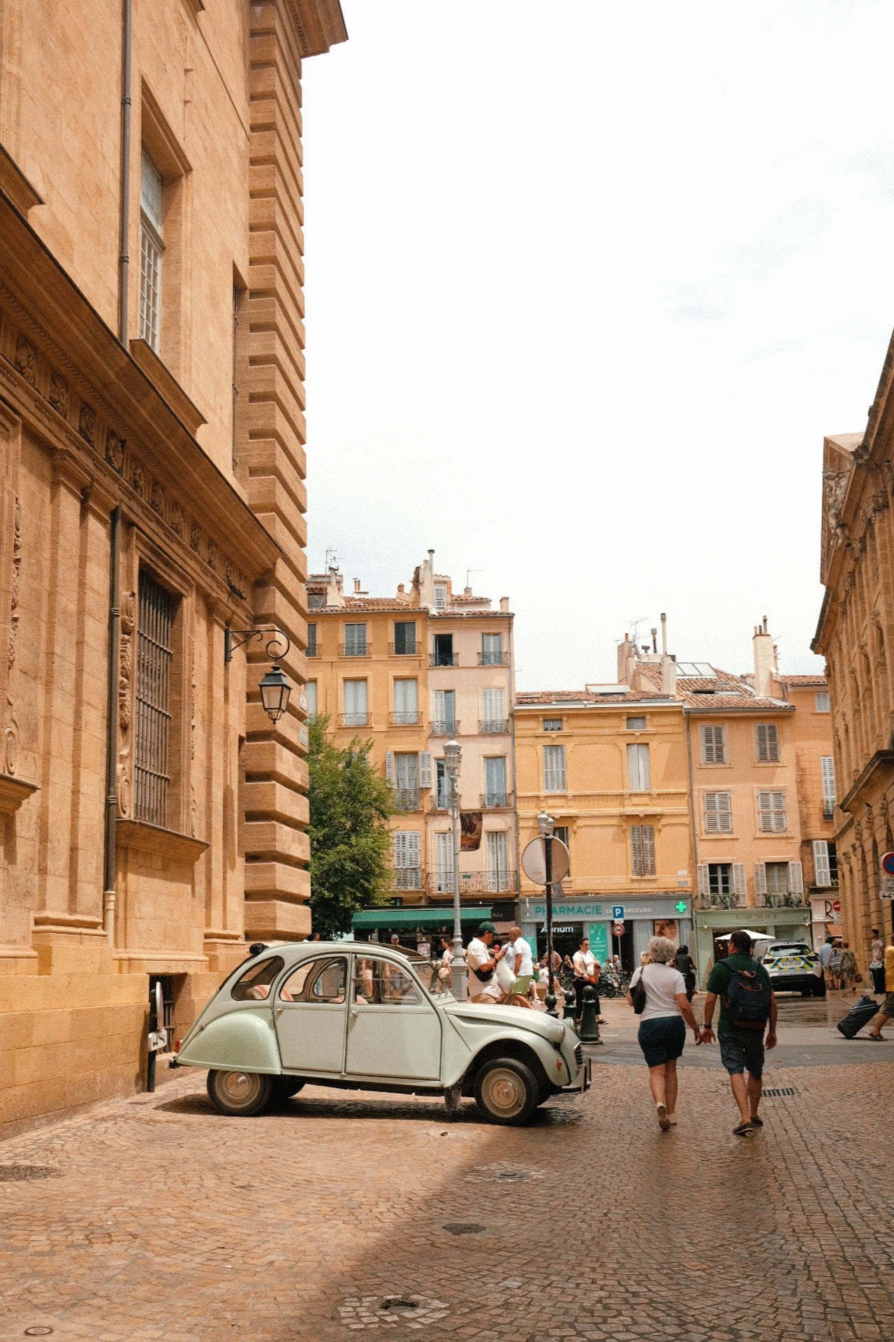 Rue pavée bordée de bâtiments en pierre beige, avec une voiture ancienne arrêtée sur le côté et des piétons marchant, certains tenant la main à Aix en Provence