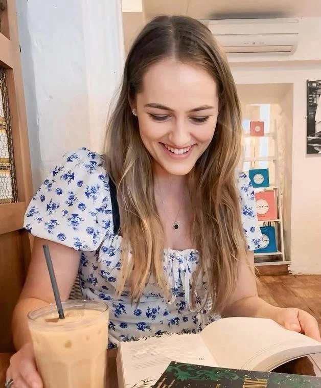 A young woman with long, wavy hair wearing a white blouse with blue floral patterns, sitting at a table in a cafe. She is smiling while looking at an open book, with a glass of iced coffee with a black straw in front of her.