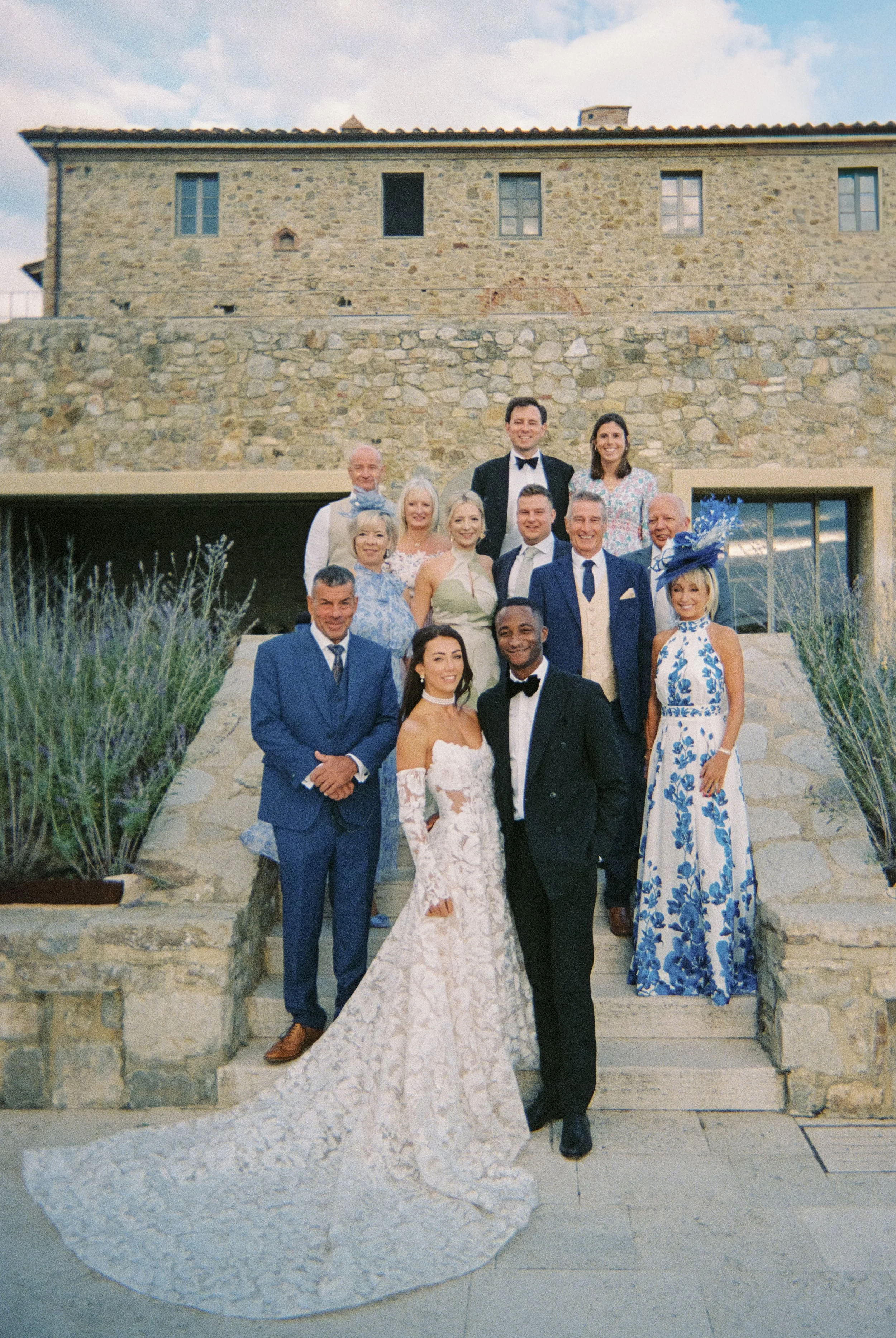 A stylish and editorial family group portrait during a Tuscany wedding in front of a rustic stone villa at podere tesoro, capturing elegant guests, soft pastel attire, and the timeless architecture of the Tuscan countryside captured on a 35mm film