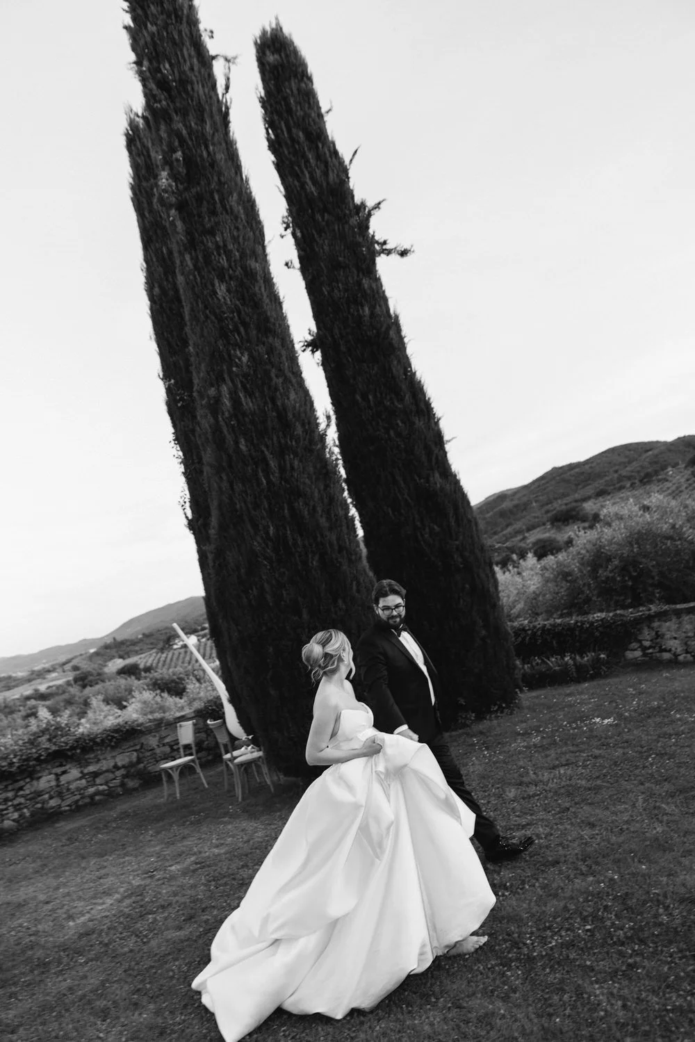 Bride and groom walking hand in hand beneath towering cypress trees at La Palagina in Tuscany for their wedding. A cinematic, black-and-white wedding photo of quiet connection