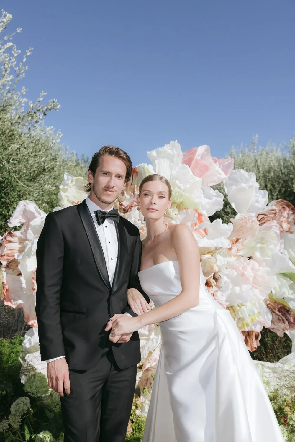 editorial-style couple portrait at a braccialieri wedding in sicily. the bride in a minimalist white Galia Lahav gown and the groom in classic hugo boss black tie, framed by an artful backdrop of oversized pastel florals under the olive trees