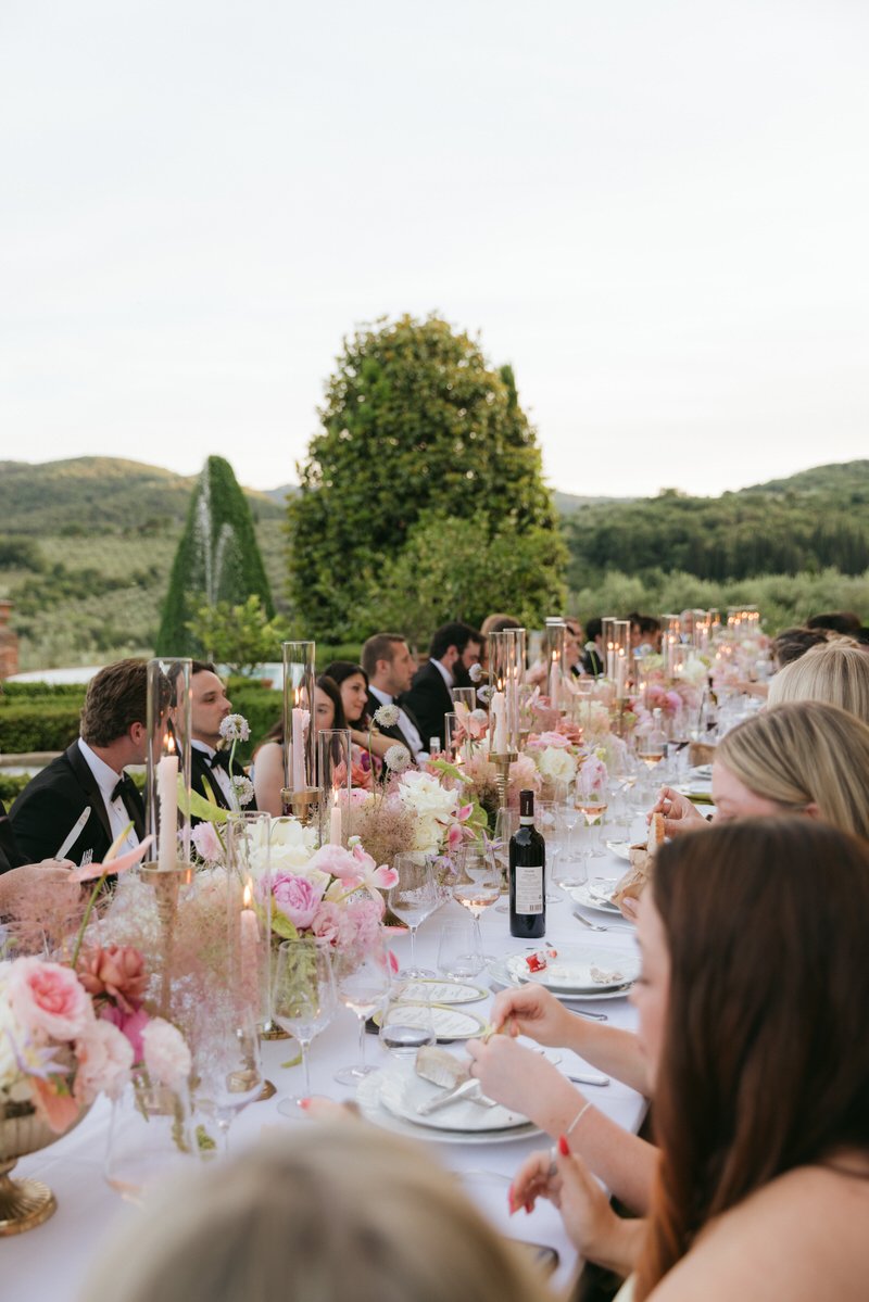 Detail of a candlelit reception table with guests and with pink florals and wine glasses at a Palagina Tuscany wedding in Italy