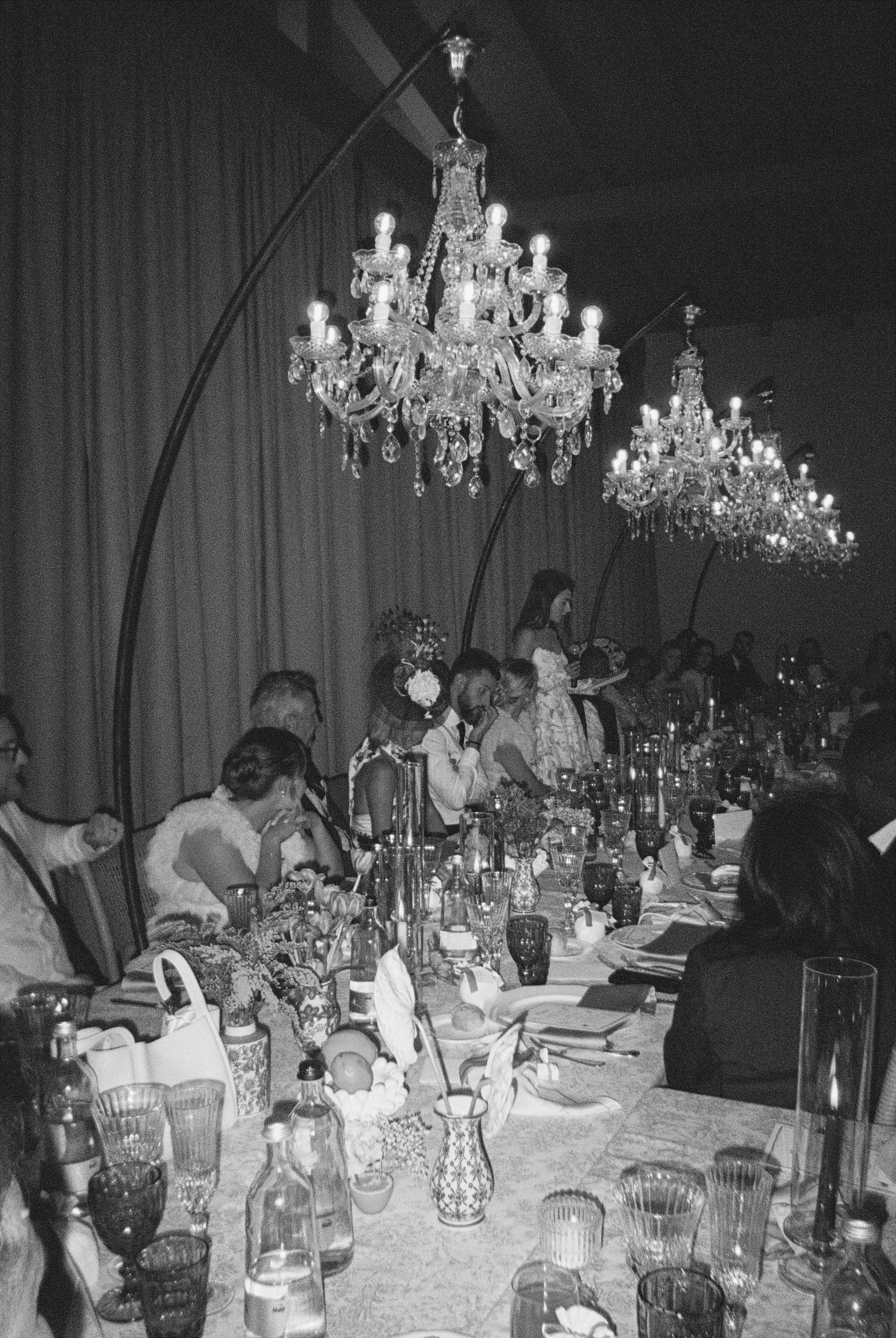 A timeless Tuscany wedding reception set beneath dramatic crystal chandeliers, with candlelit tables, layered textures, and elegant guests gathered for a wedding dinner in an elegant Tuscan venue at podere tesoro captured on a 35mm film