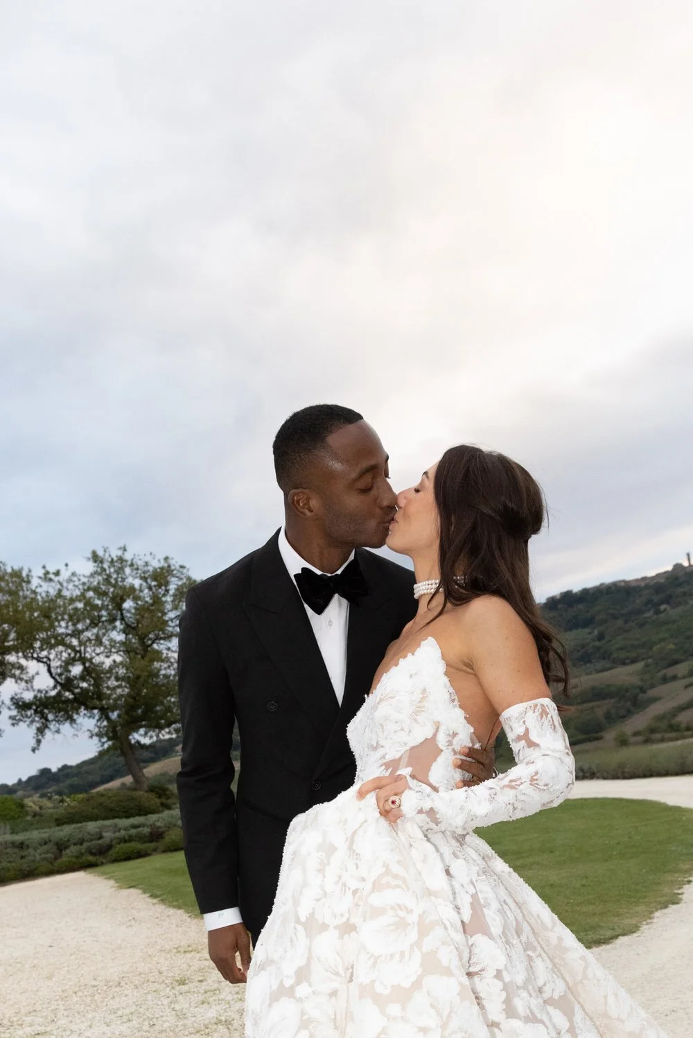 A romantic outdoor kiss framed by soft hills and natural light, blending editorial elegance with genuine feeling. A signature La Foce Tuscany wedding portrait with soulful presence