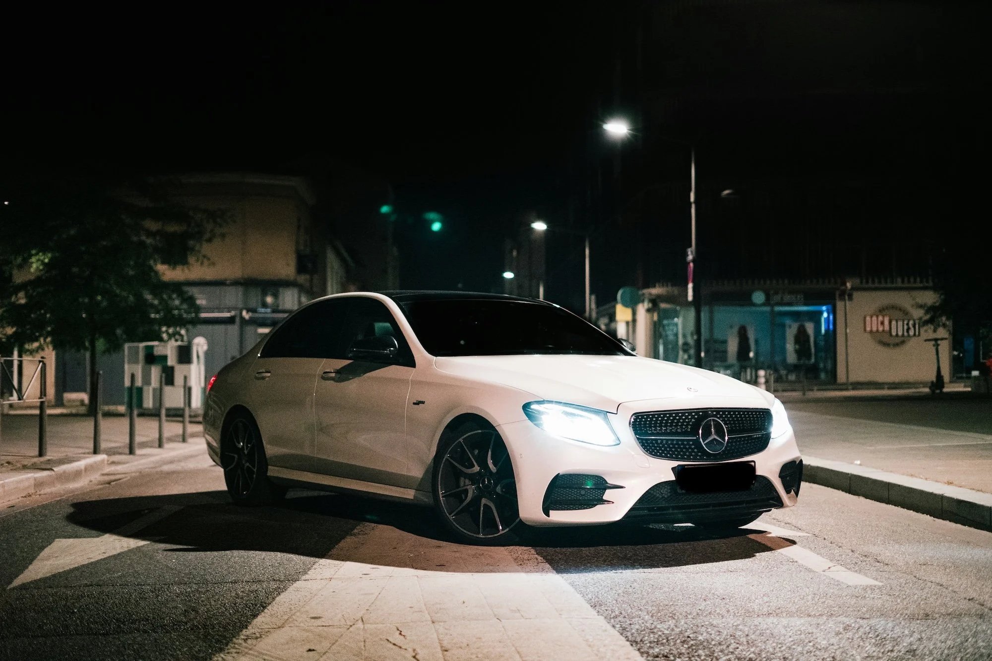 A white Mercedes-Benz coupe parked at night on a city street with illuminated headlamps, streetlights, and storefronts in the background.