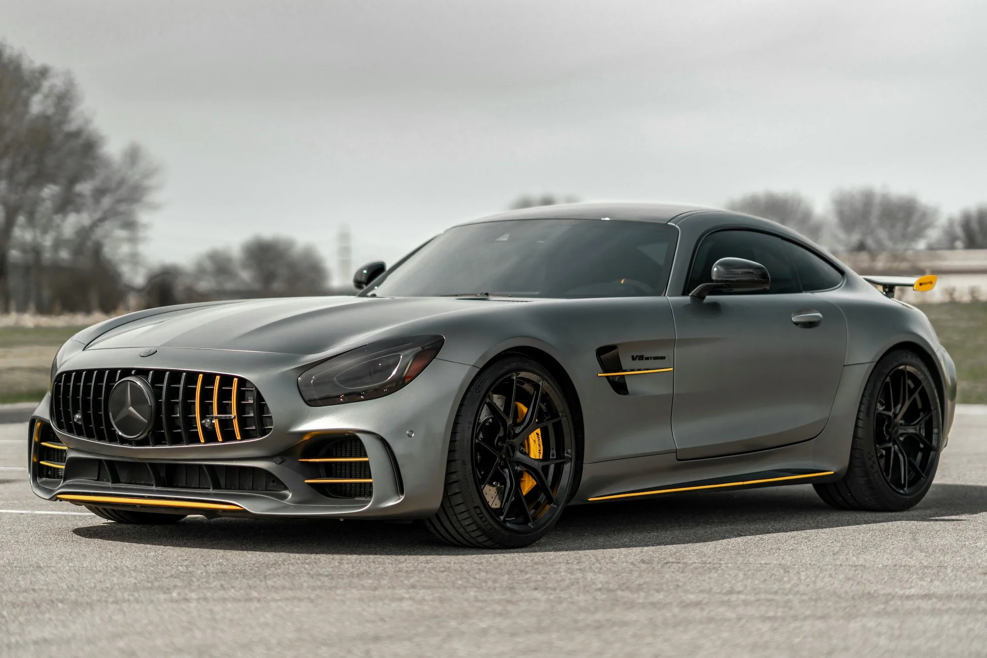 A silver Mercedes-Benz sports car with black and yellow accents parked on an empty road under an overcast sky.