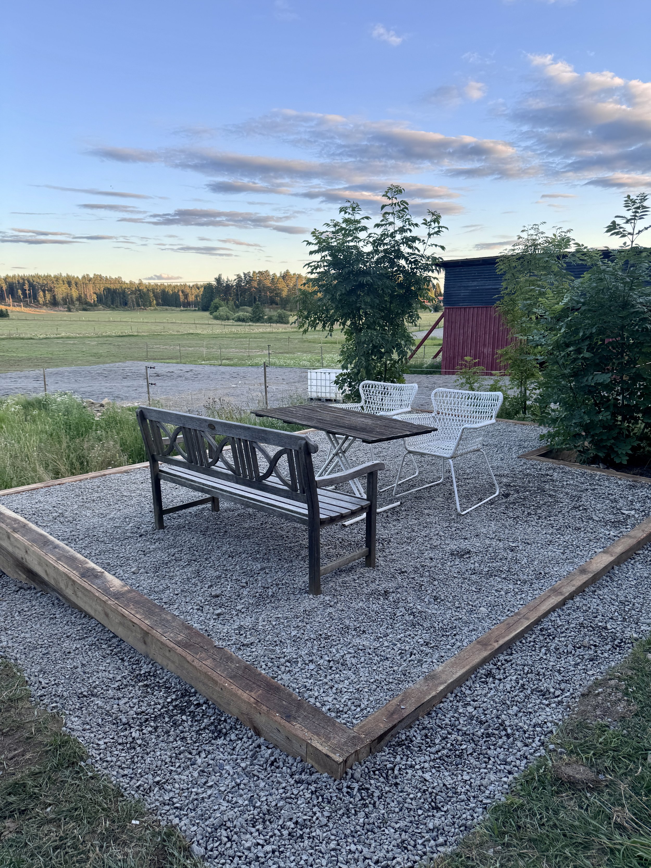 A gravel patio with outdoor furniture, including a wooden bench, white metal chairs, and a wooden table, surrounded by green grass and trees, with a red barn and open field in the background, under a partly cloudy sky.