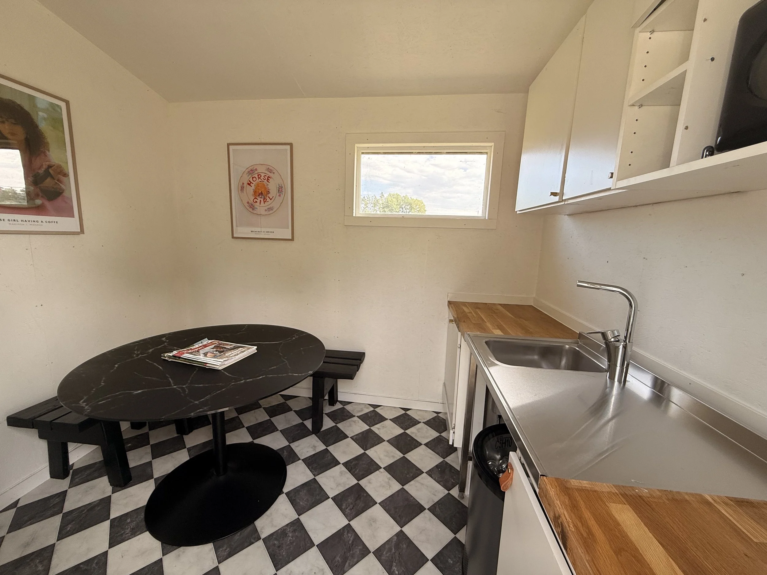 A small kitchen area with a black marble round table, a black bench, a white kitchen cabinet with an open shelf, a stainless steel sink, a window showing trees outside, and framed posters on the wall, one with a girl and the other with a horse.