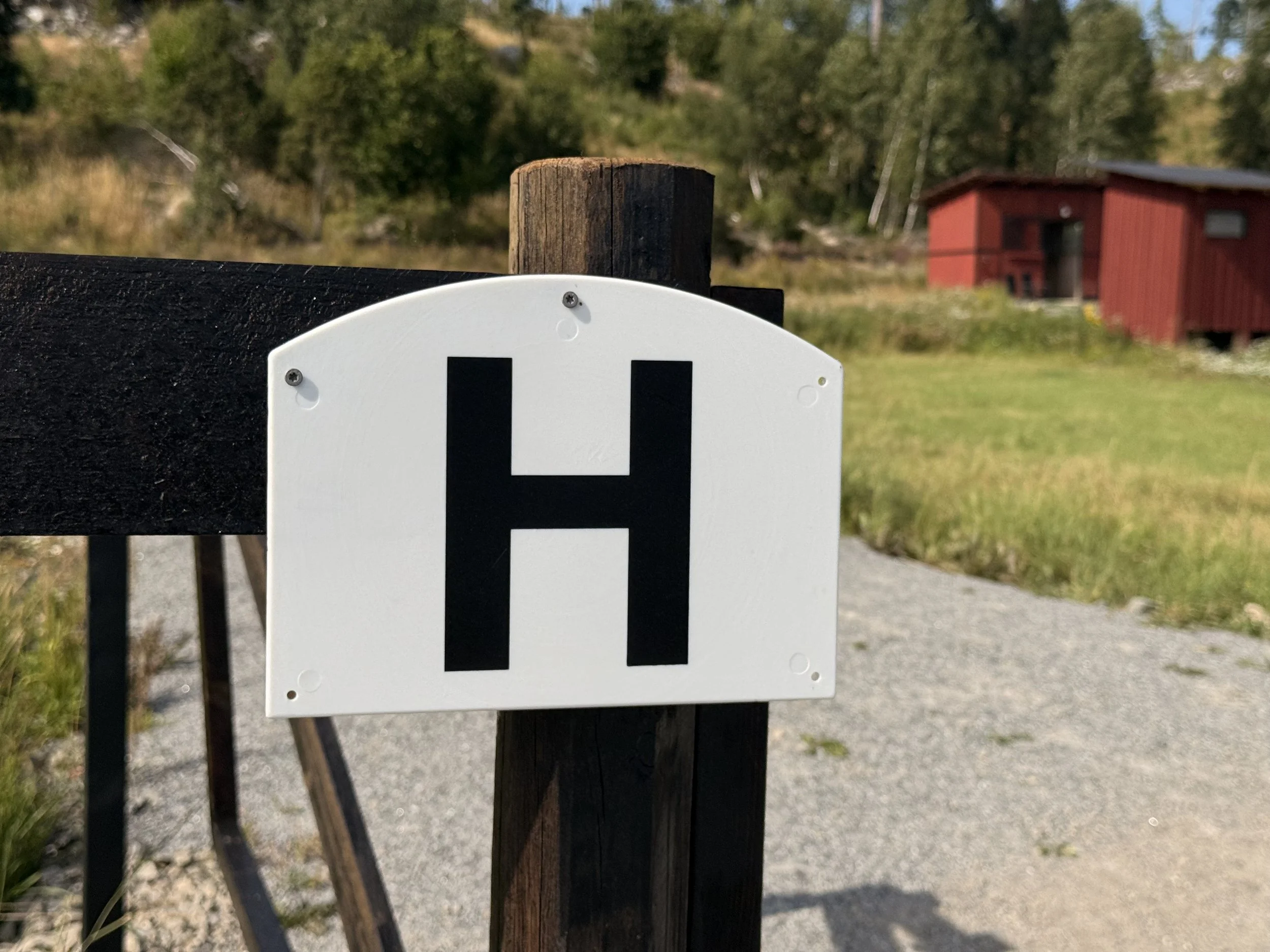 Close-up of a white sign attached to a black wooden post with black lettering, with a blurry background of greenery, a red building, and yellow flowers.
