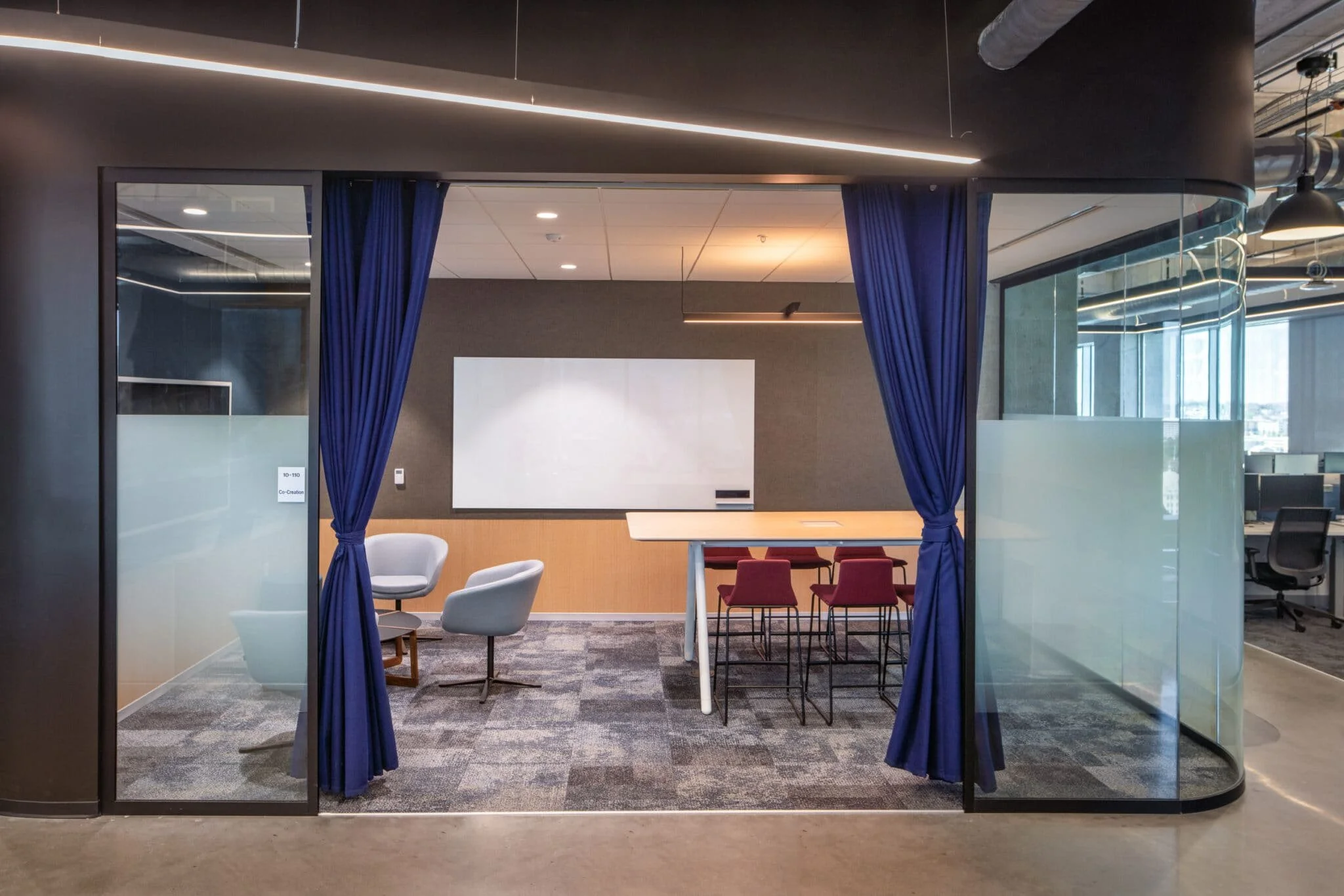 Glass-front co-creation room with blue curtains, whiteboard, lounge chairs, and a high worktable.